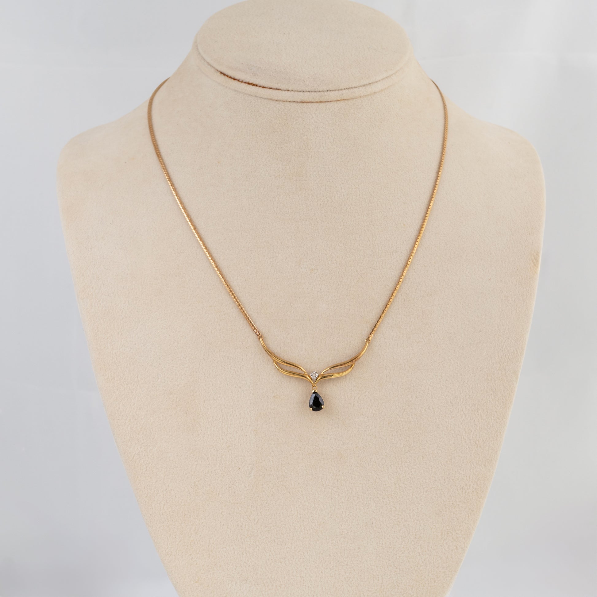 A delicate gold chain necklace with a small teardrop-shaped dark gemstone pendant, displayed on a beige fabric jewelry bust against a light gray background.