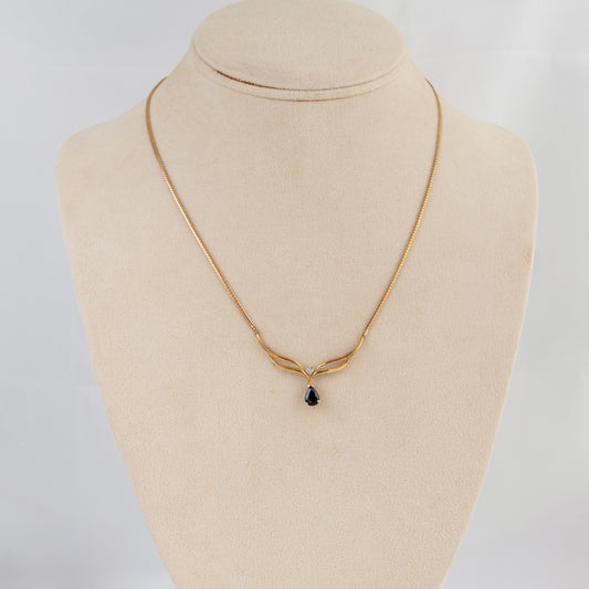 A delicate gold chain necklace with a small teardrop-shaped dark gemstone pendant, displayed on a beige fabric jewelry bust against a light gray background.