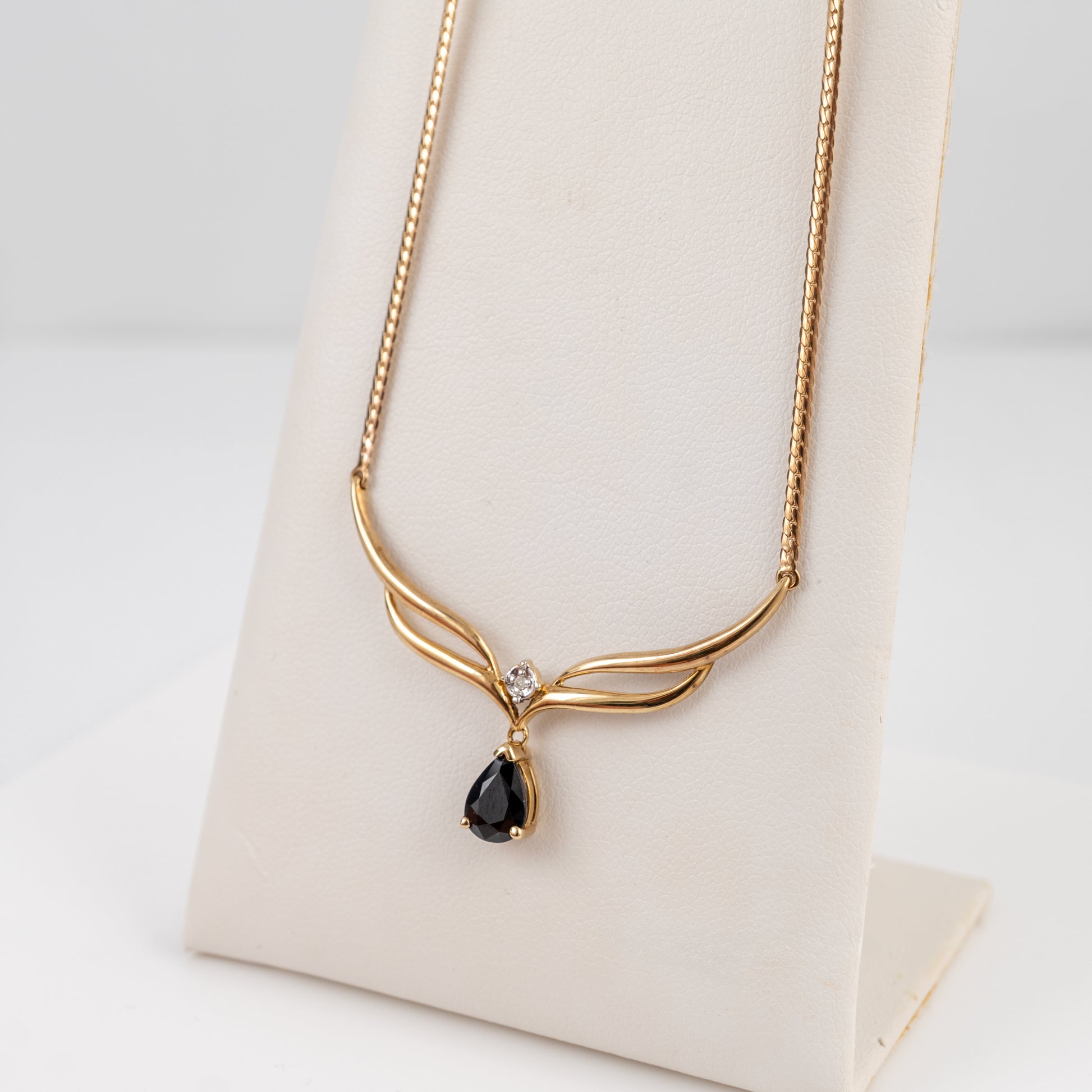 A gold necklace with a curved openwork pendant and a teardrop-shaped black gemstone, displayed on a cream-colored jewelry stand against a white background.