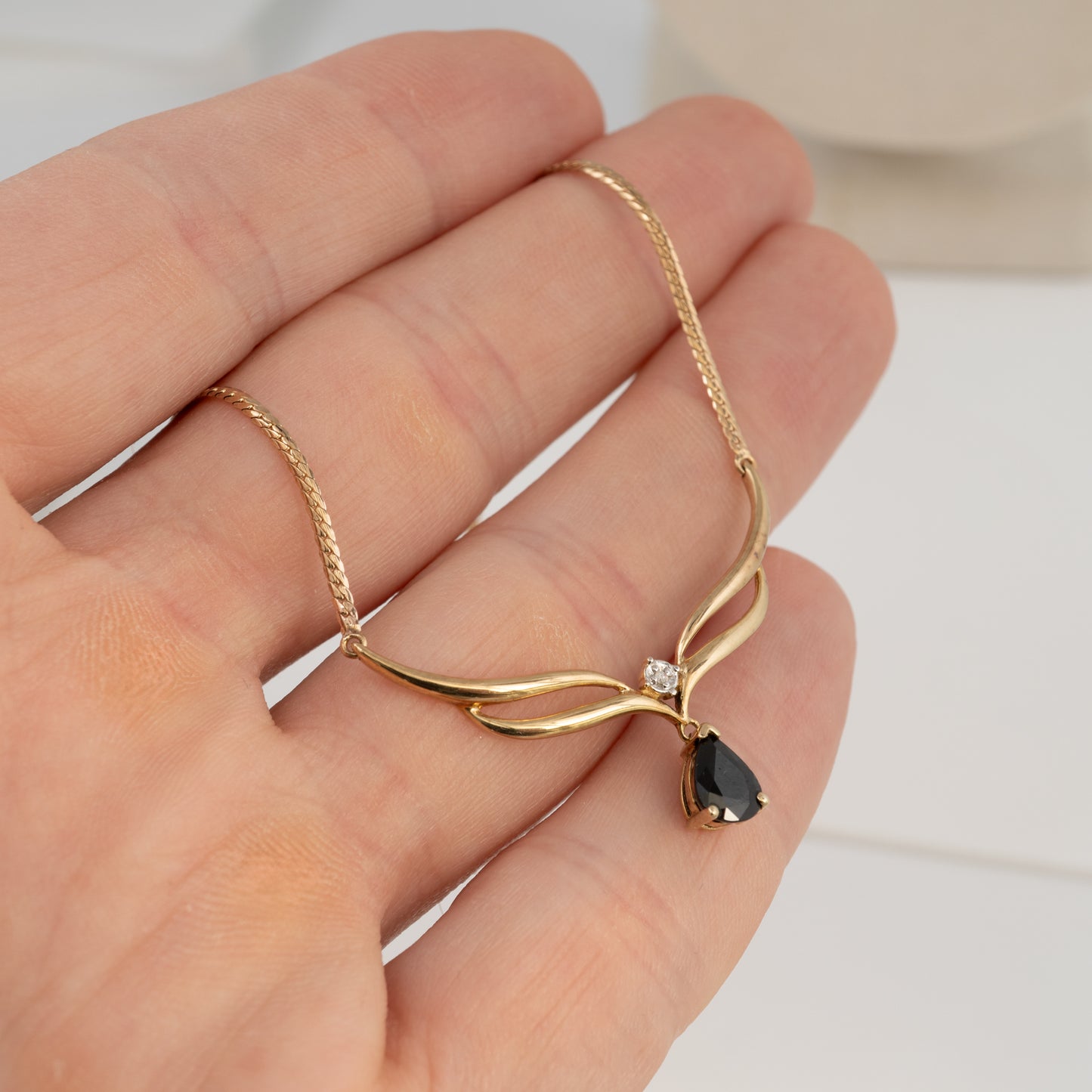 A gold-toned necklace with a curved branch-like pendant, a small clear stone, and a dark teardrop gemstone, displayed on a person's hand.