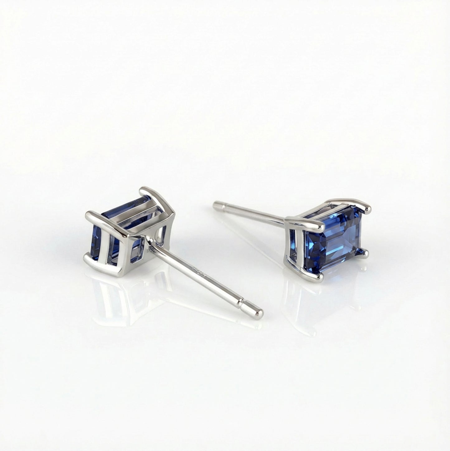 A pair of silver stud earrings with rectangular blue gemstones, displayed on a clean white reflective surface.