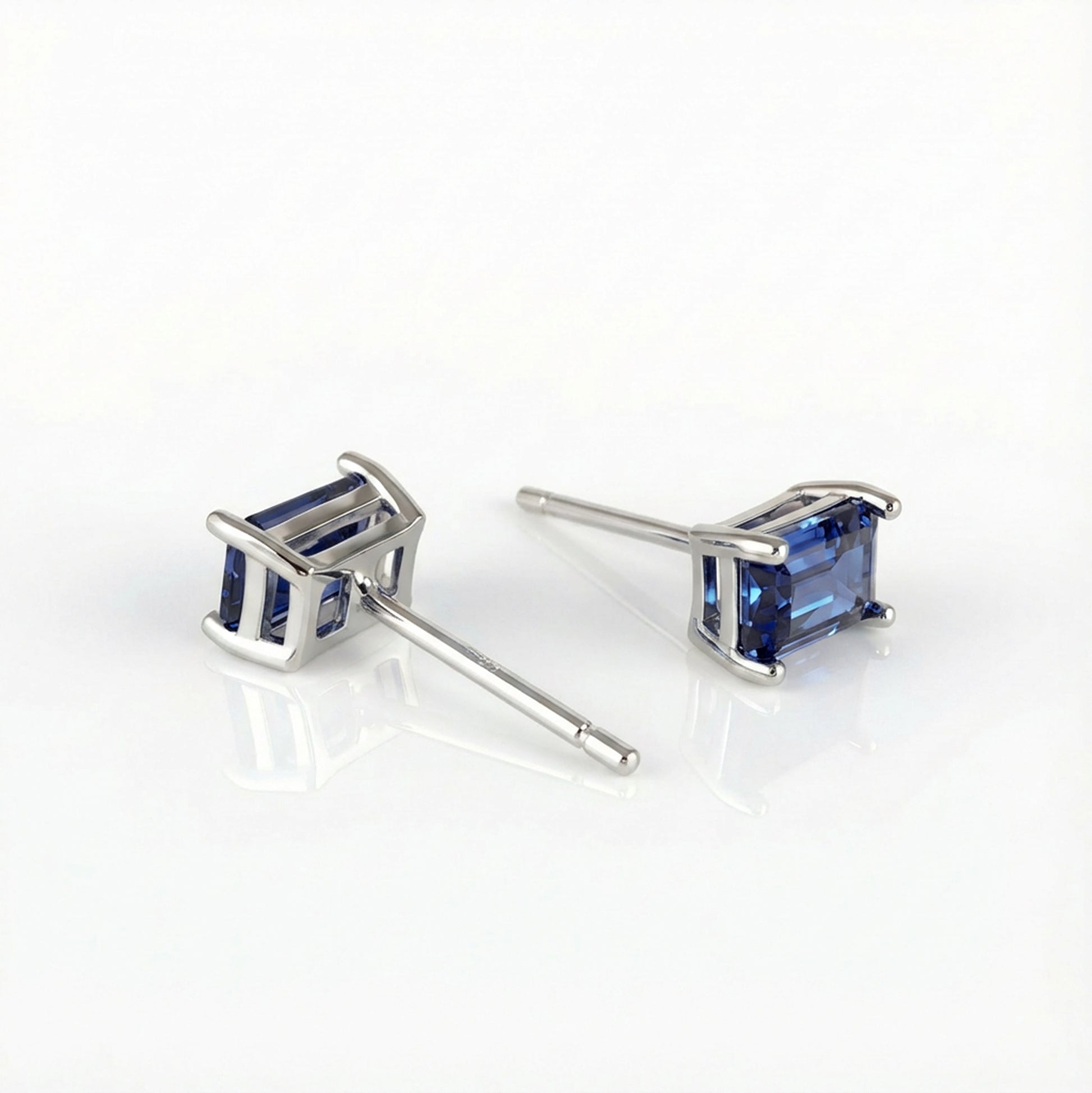 A pair of silver stud earrings with rectangular blue gemstones, displayed on a clean white reflective surface.