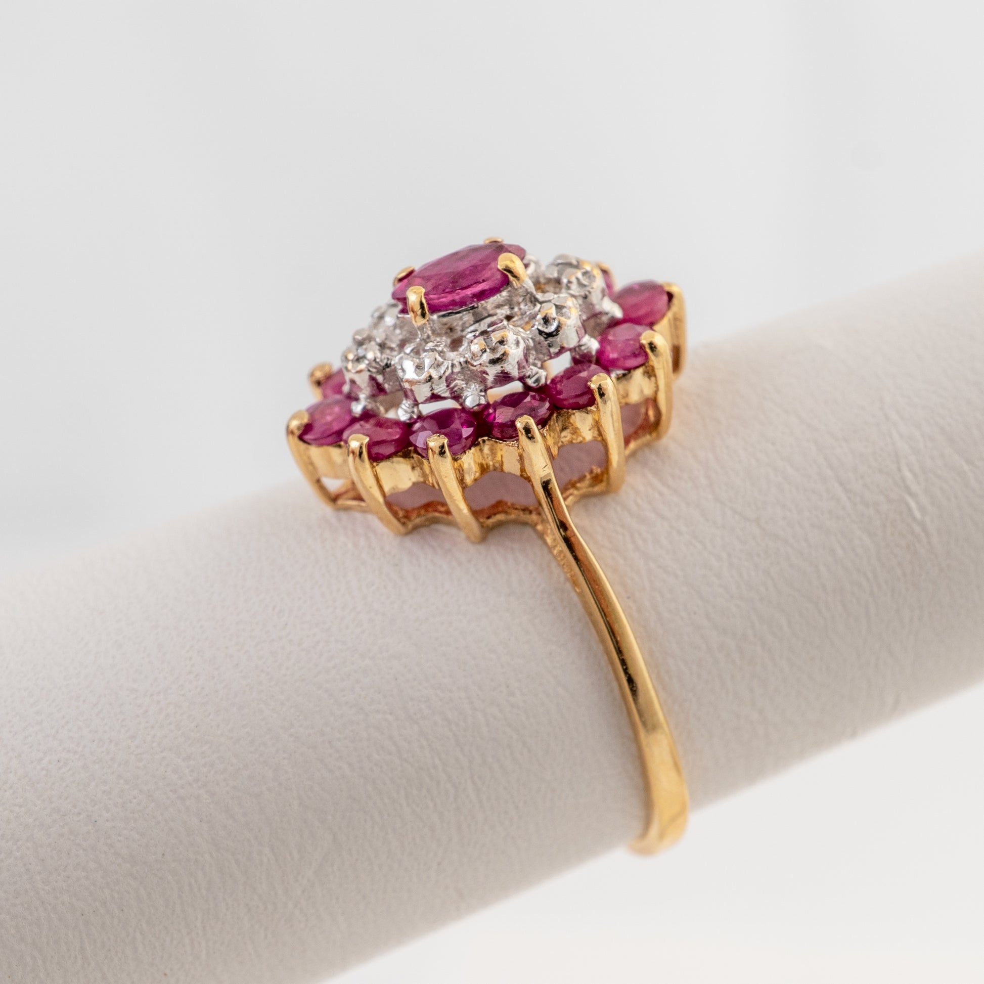 A yellow gold ring with a floral cluster setting featuring pink gemstones and clear stones, displayed on a white ring stand against a light gray background.