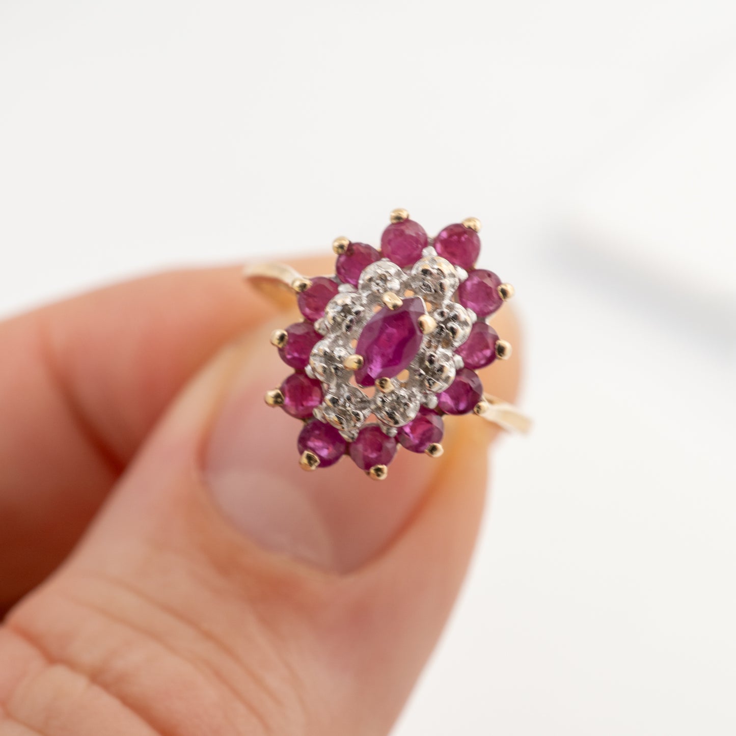 A close-up of a gold ring with a floral cluster setting featuring a central oval ruby and surrounding round rubies and small clear stones, shown being held between fingers against a soft white background.