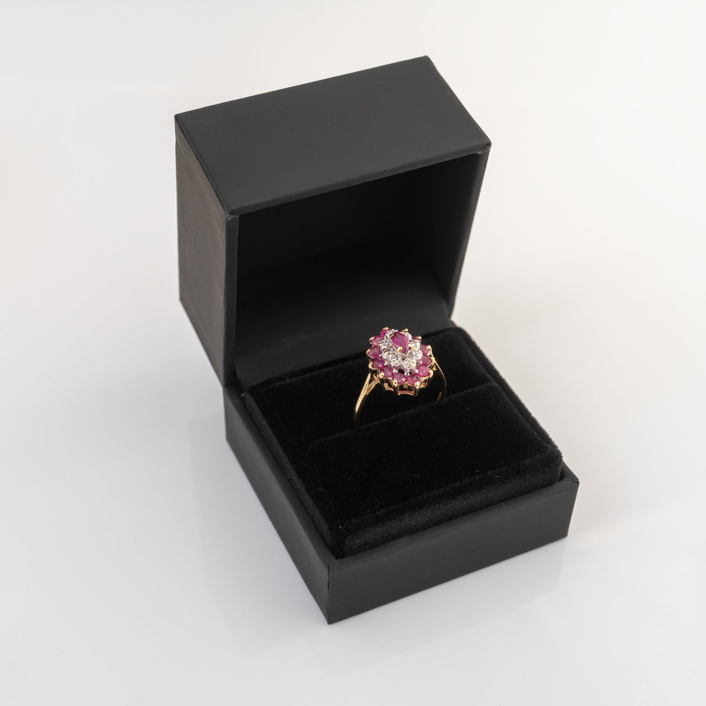 A yellow gold ring with a pink gemstone cluster setting, presented inside an open black jewelry box on a white background.