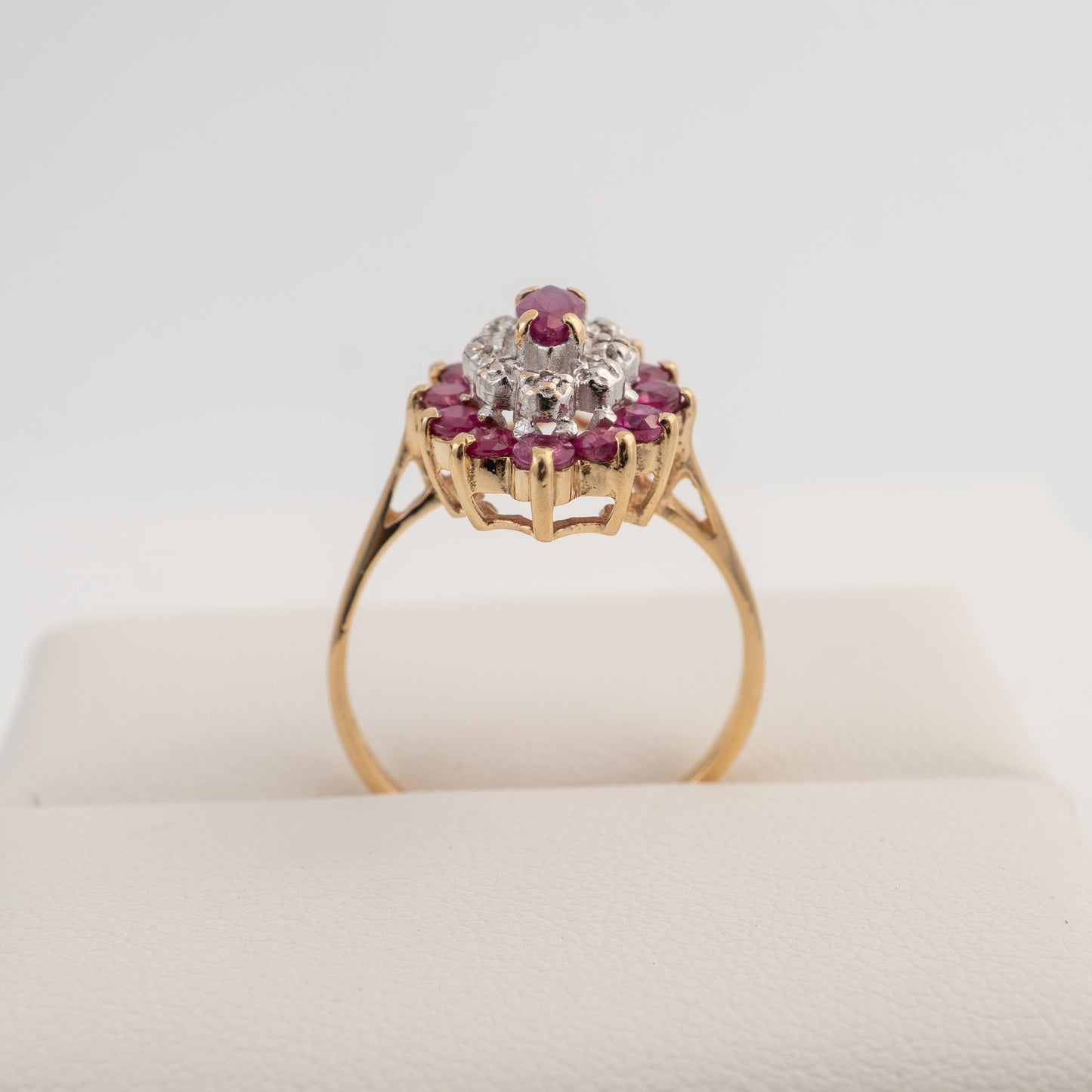 A yellow gold ring with a floral cluster setting featuring a central clear gemstone surrounded by small pinkish-red round stones, displayed on a cream-colored ring stand against a white background.