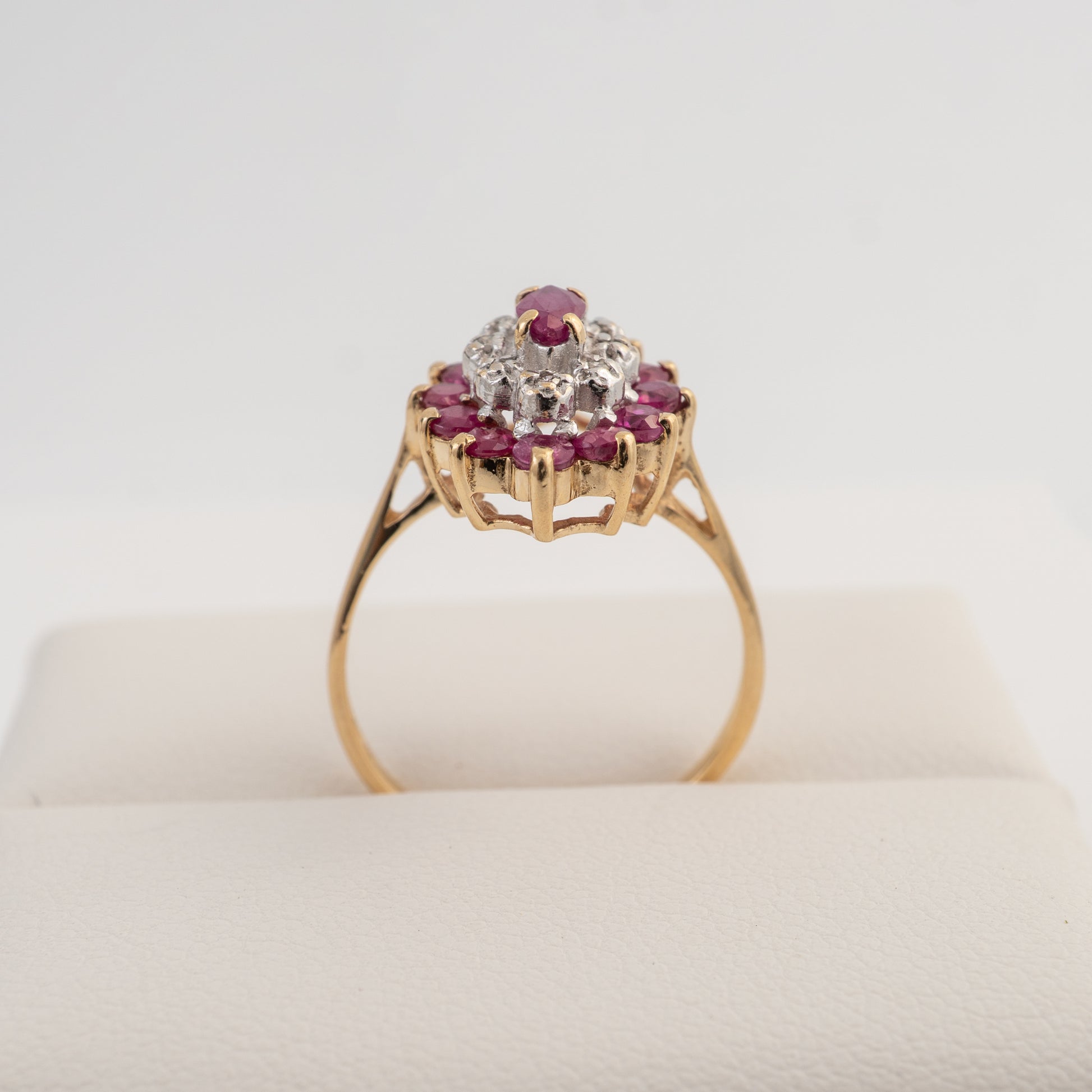 A yellow gold ring with a floral cluster setting featuring a central clear gemstone surrounded by small pinkish-red round stones, displayed on a cream-colored ring stand against a white background.