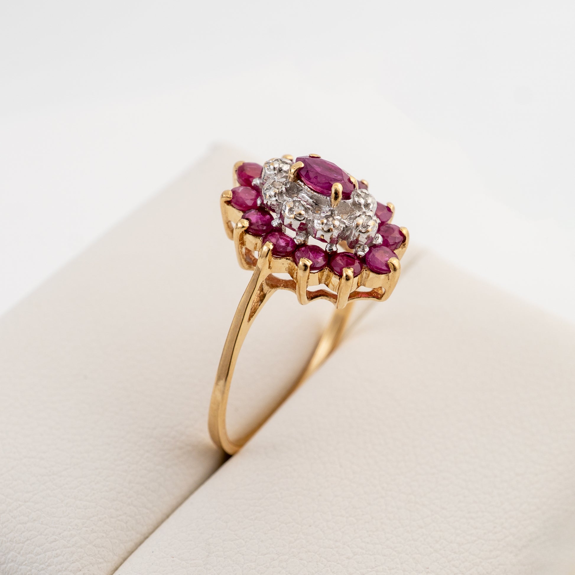 A yellow gold ring featuring a floral cluster setting with a central cluster of clear stones surrounded by a halo of round pinkish-red gemstones, displayed on a cream-colored jewelry stand.