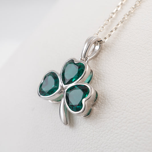 A silver shamrock pendant necklace with three heart-shaped dark green crystal stones, resting on a textured white surface.