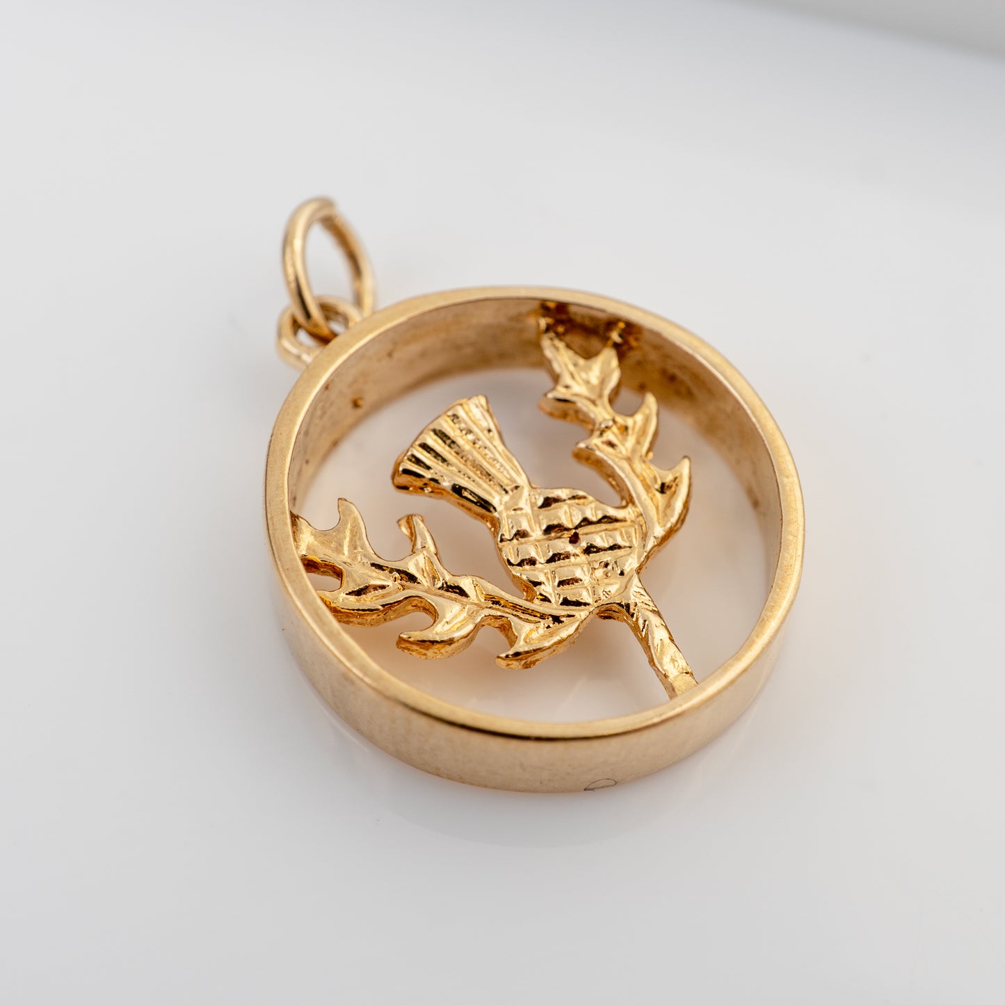 A gold pendant featuring a textured thistle inside an open circular frame, shown on a plain light background.