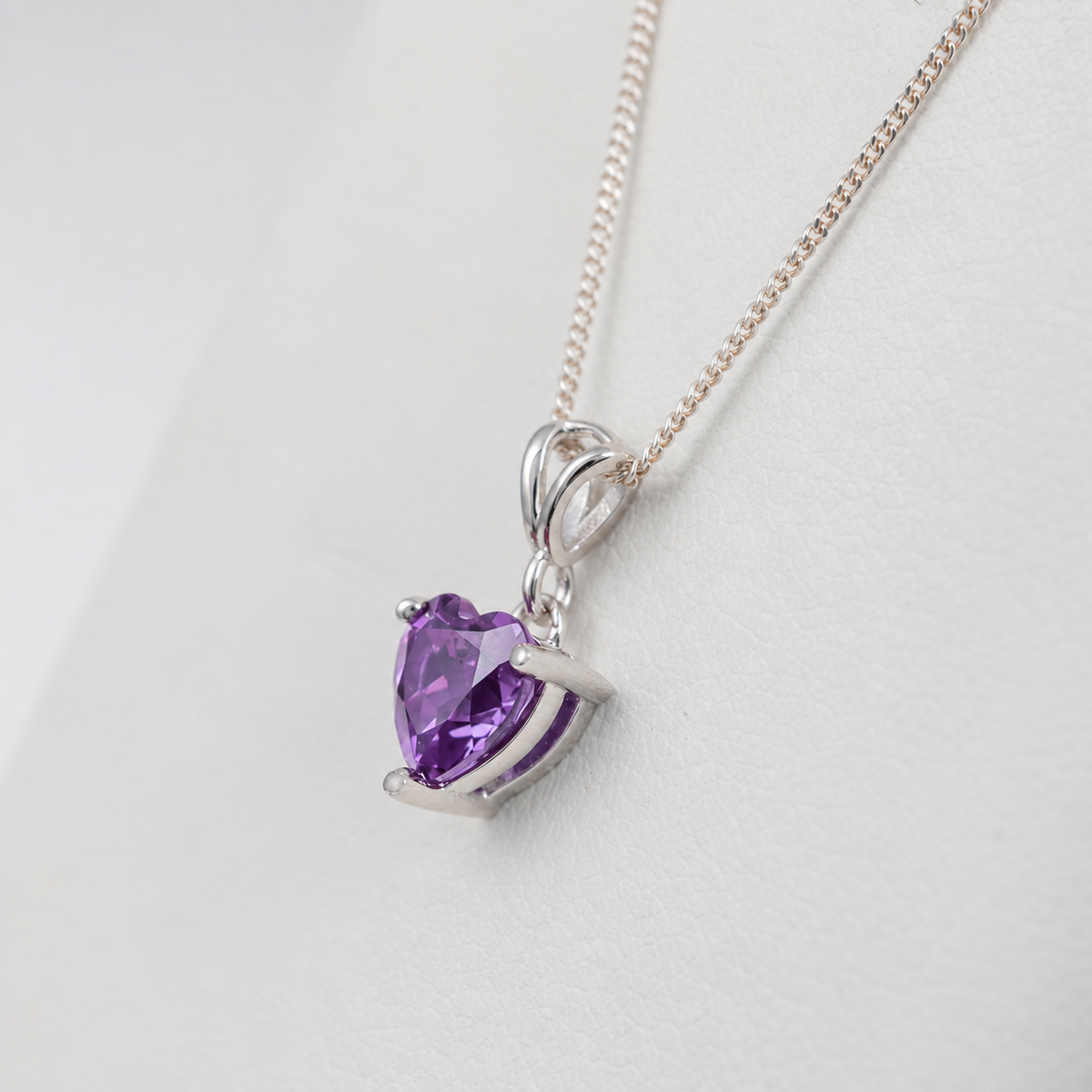 A heart-shaped amethyst pendant in a silver-colored setting on a delicate silver chain, displayed on a white background.