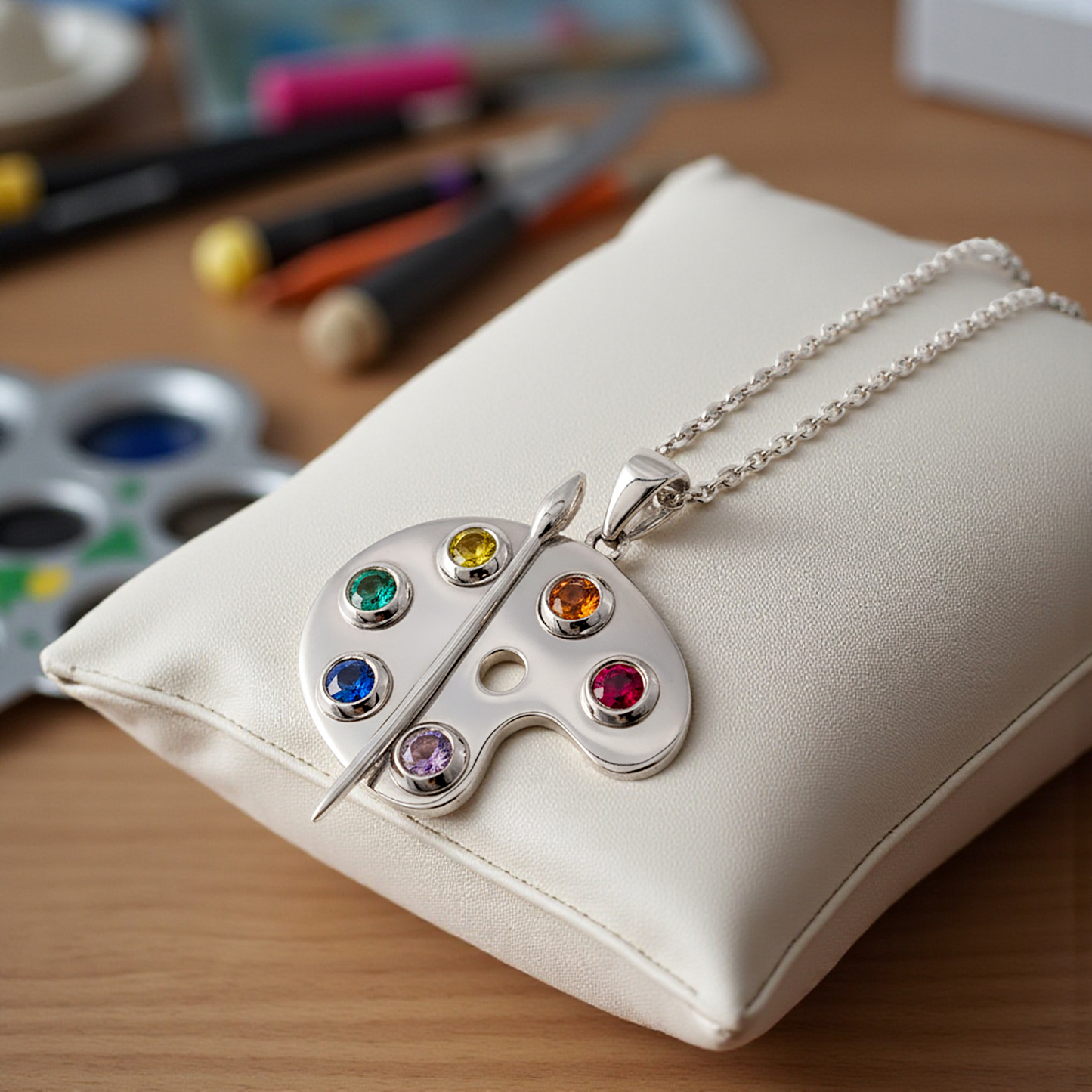 Silver necklace with a colorful gemstone palette pendant on a white cushion, with art supplies in the background.