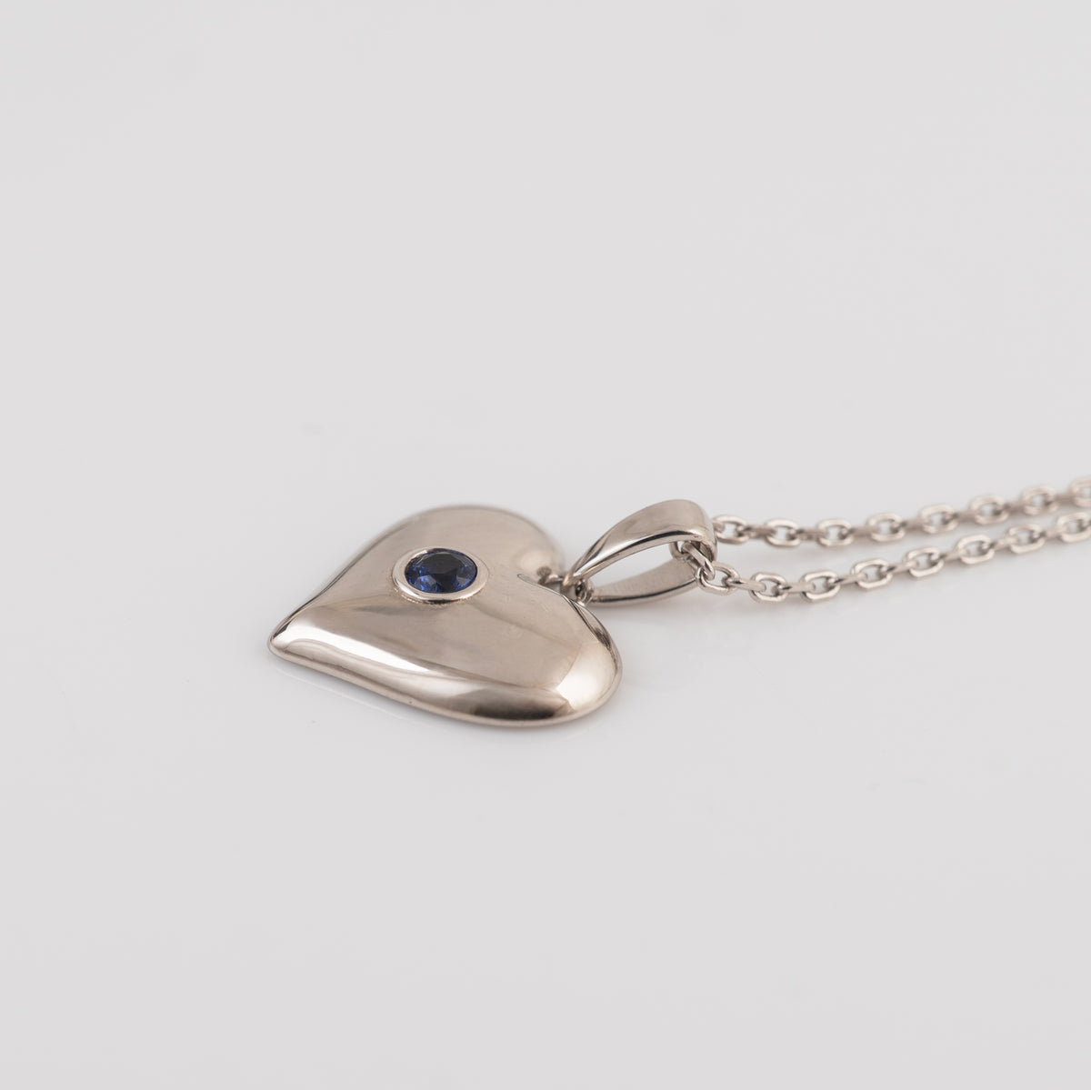 A polished silver heart-shaped pendant necklace with a small round blue sapphire gemstone inset, shown on a matching silver chain against a light gray background.