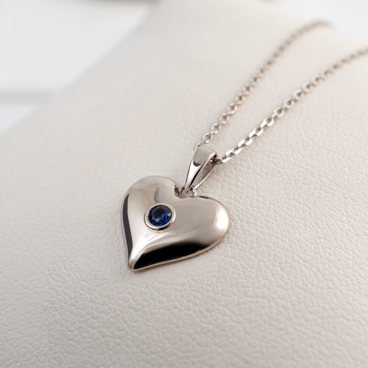 A polished silver heart pendant necklace with a small round blue sapphire gemstone, resting on a textured white display stand.