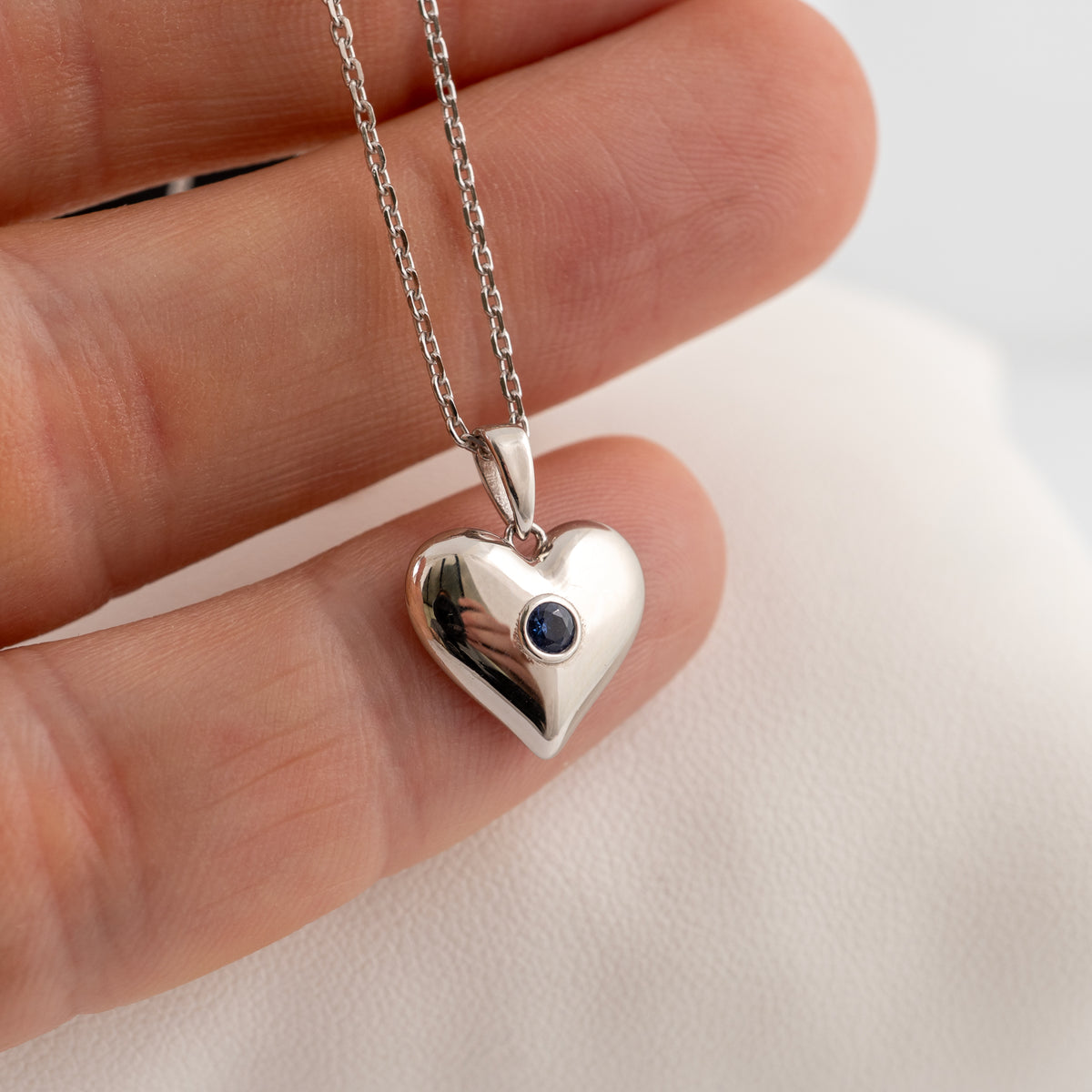 Silver heart pendant with a round lab-grown blue sapphire held against fingers to show its size