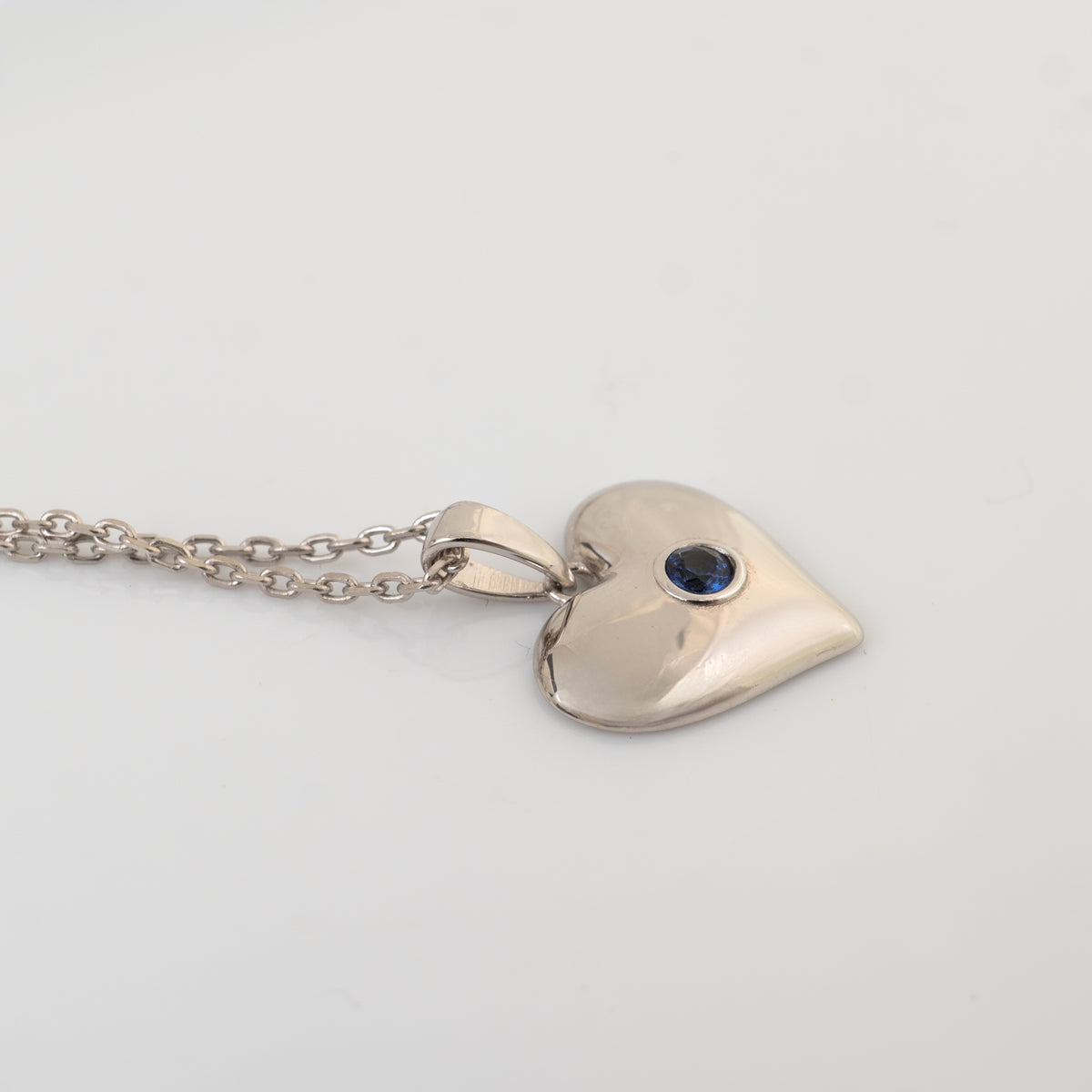 A polished silver heart pendant necklace with a small round dark blue sapphire gemstone set in the center, displayed on a clean white background.