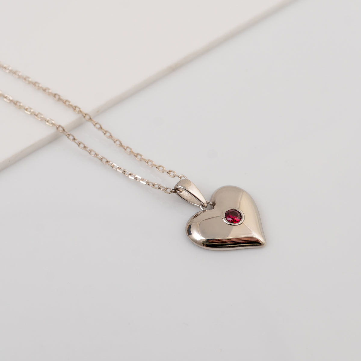 A silver heart pendant necklace with a small lab grown ruby gemstone accent, displayed on a clean white background.