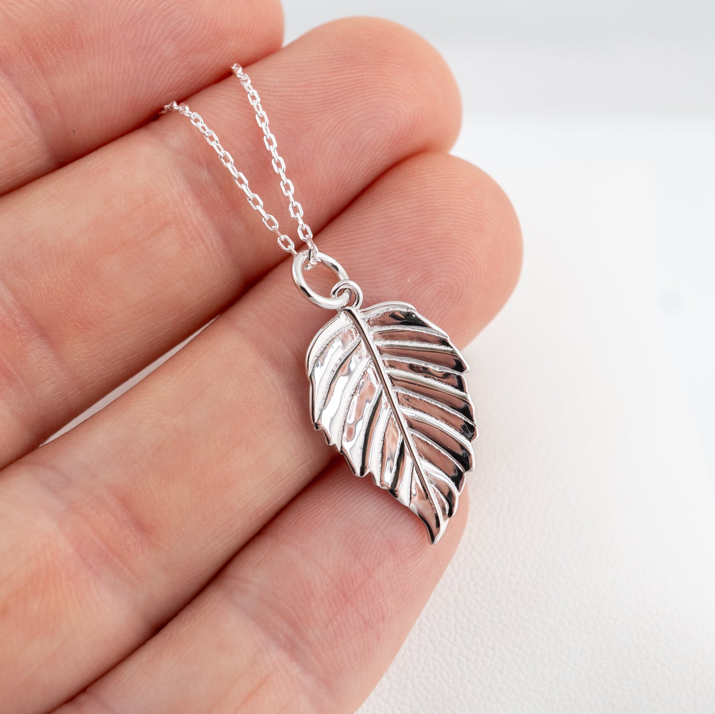 Silver leaf necklace held between fingers against a white background