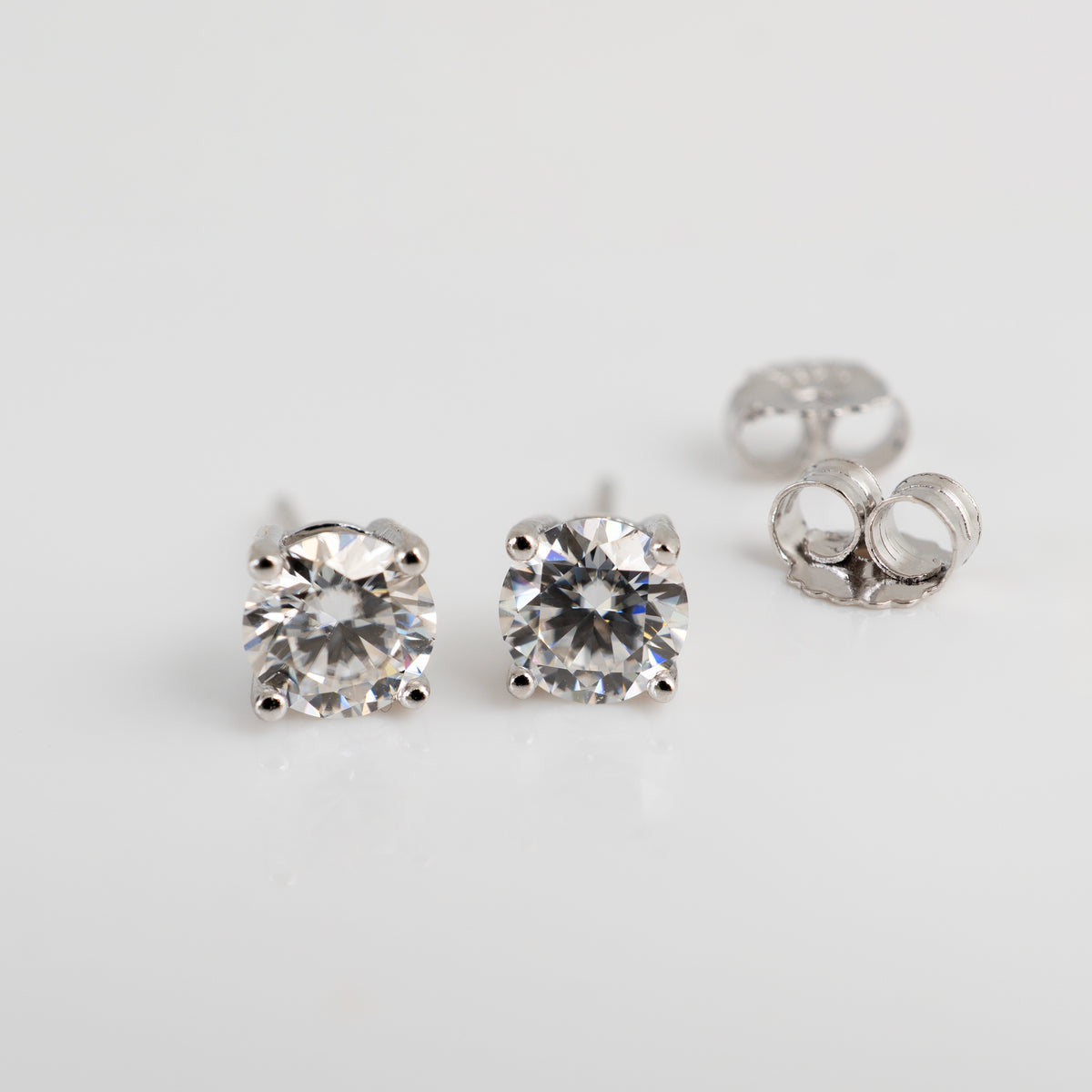A pair of round-cut moissanite solitaire stud earrings in silver-toned metal with a third earring showing the butterfly back, photographed on a clean white background.