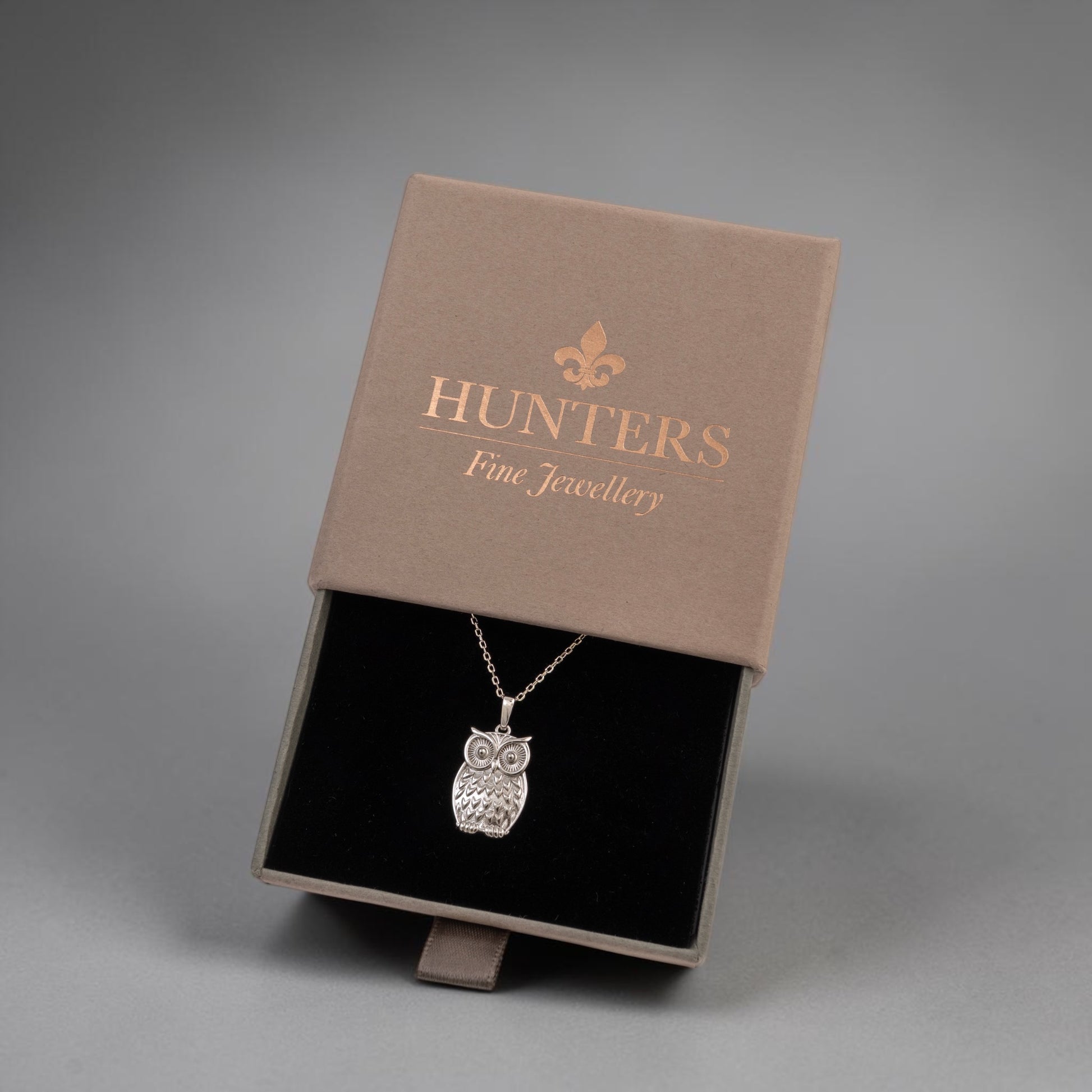A silver owl pendant necklace displayed inside an open brown presentation box with a black velvet insert. The box lid is printed with the brand name 'HUNTERS Fine Jewellery' in gold.
