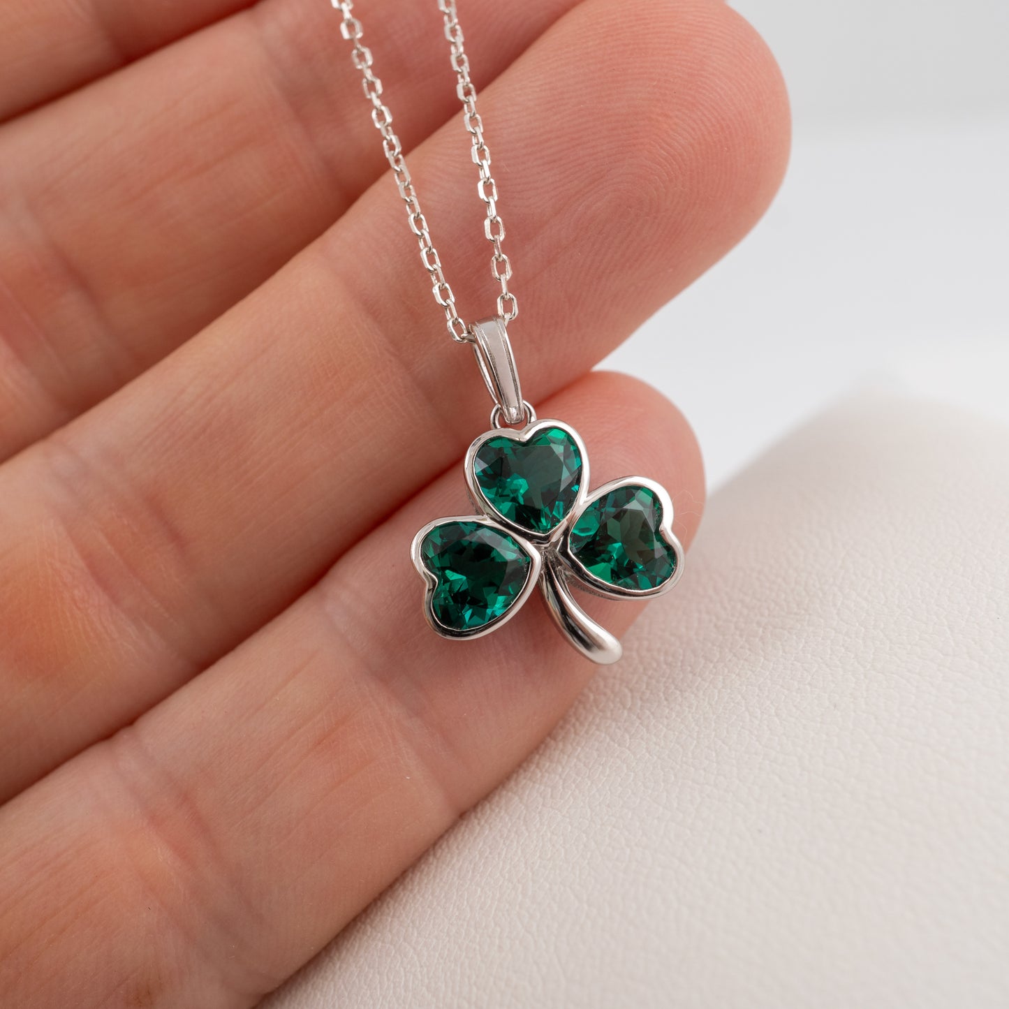 A silver shamrock pendant necklace with three heart-shaped green gemstones, shown resting on a person's fingers against a light background.