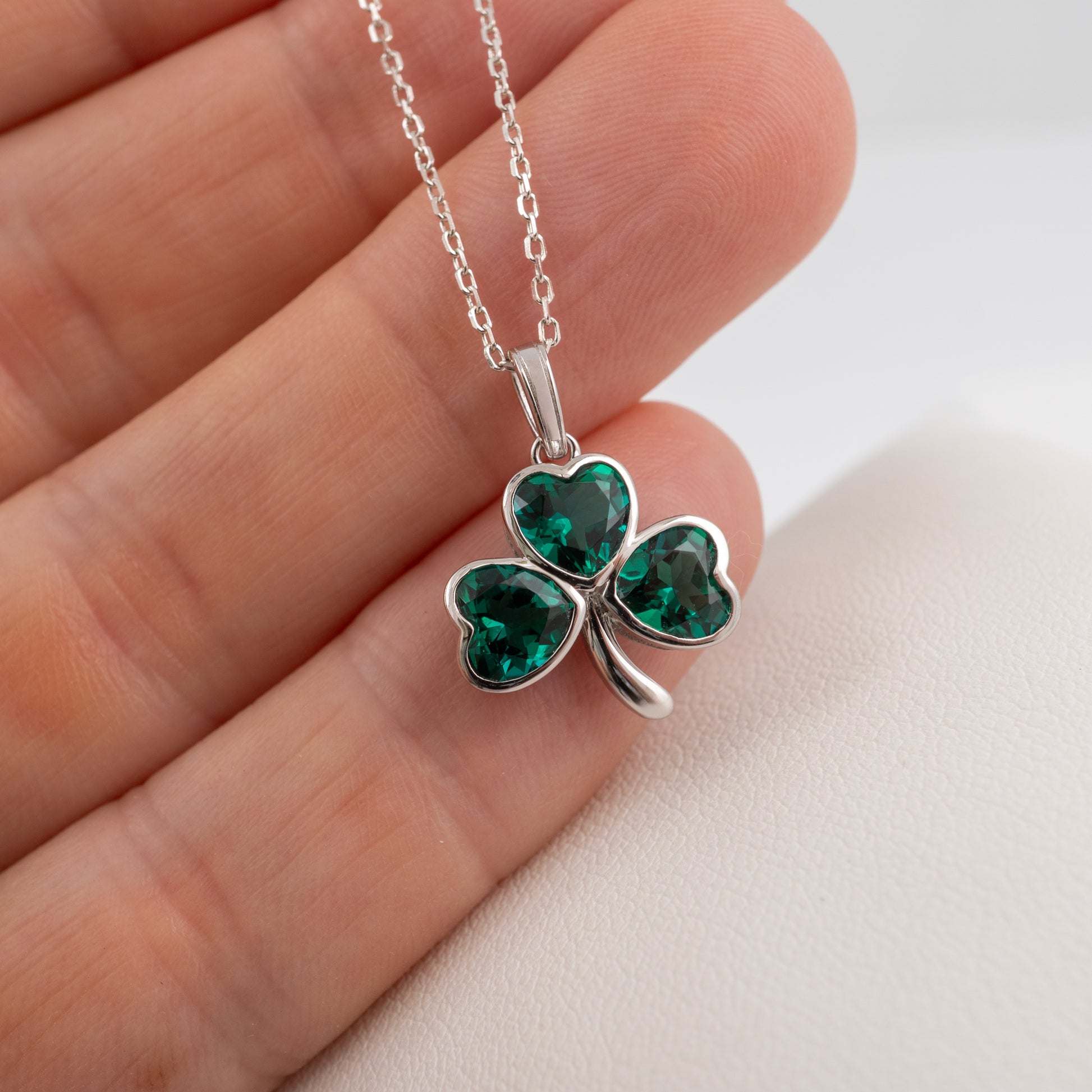A silver shamrock pendant necklace with three heart-shaped green gemstones, shown resting on a person's fingers against a light background.