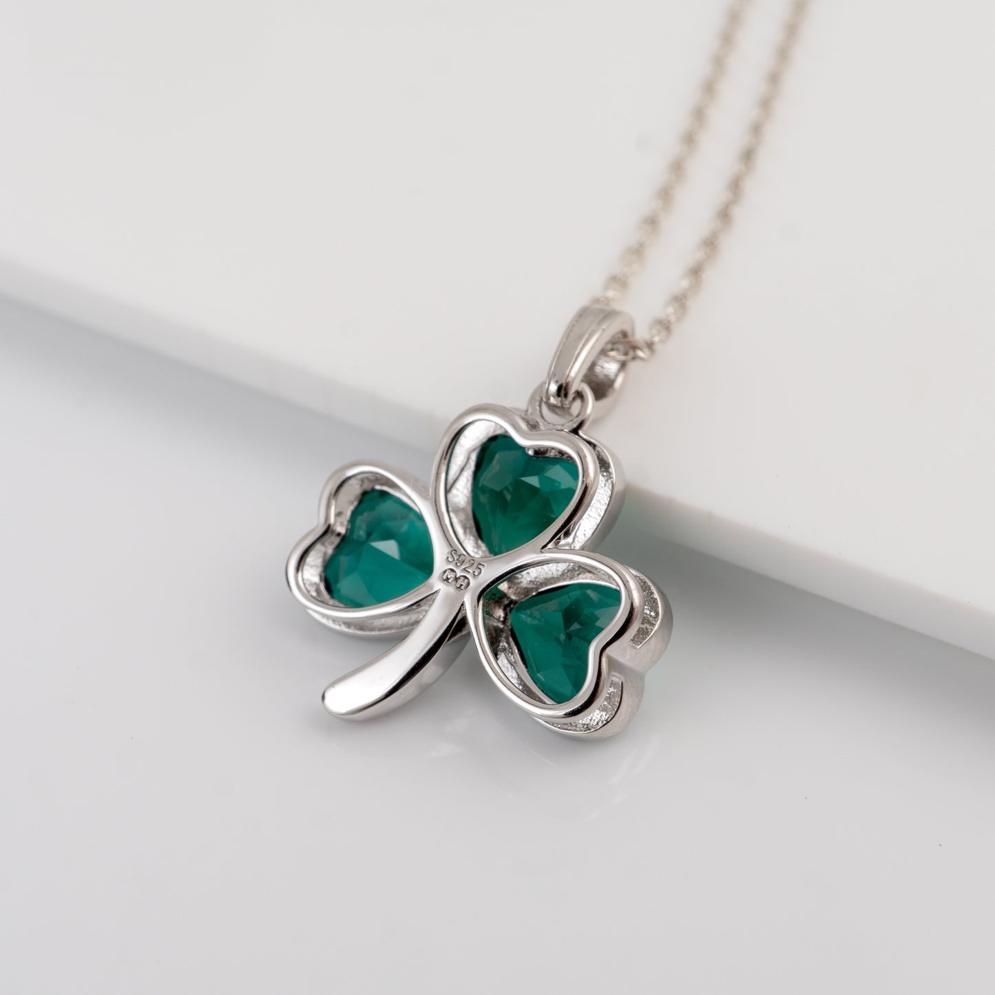 A silver shamrock pendant necklace with three heart-shaped green gemstones, resting on a clean white surface.