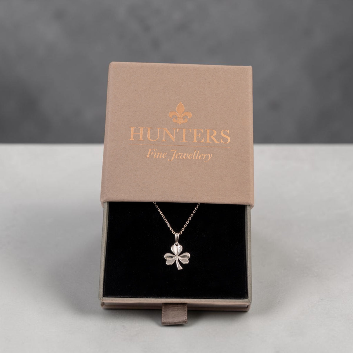 A silver shamrock pendant necklace displayed inside an open brown presentation box with the brand name 'HUNTERS Fine Jewellery' printed on the lid, set against a neutral gray background.