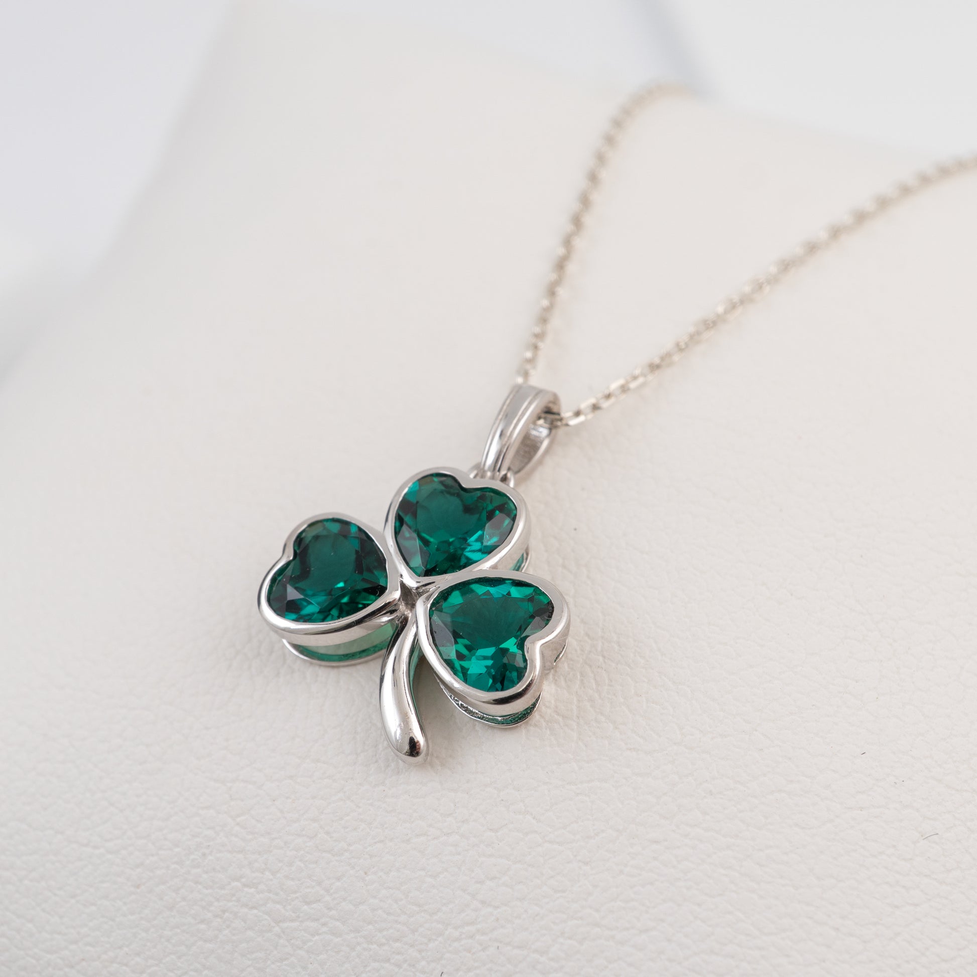 A silver shamrock pendant necklace with three heart-shaped green gemstones, displayed on a white textured surface.