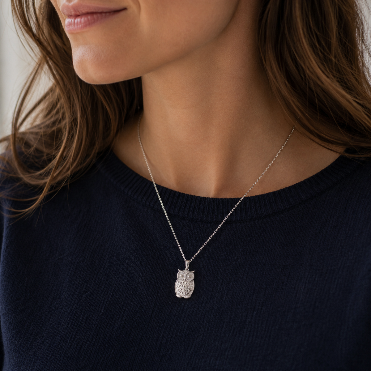 A close-up of a woman wearing a silver owl pendant necklace on a delicate chain, paired with a dark navy blue knit top.