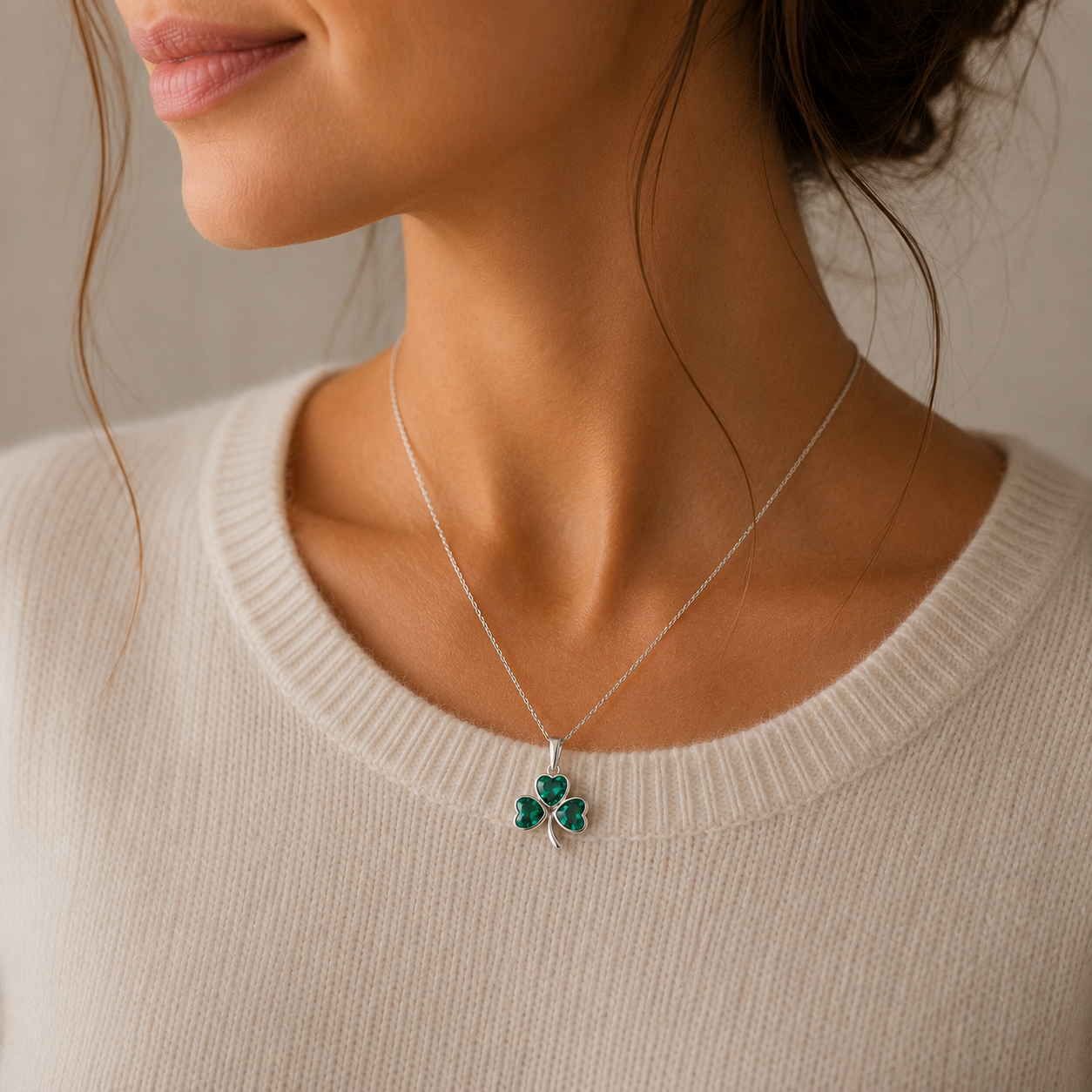A close-up of a woman wearing a delicate silver necklace with a green shamrock pendant, paired with a cream-colored ribbed knit sweater.