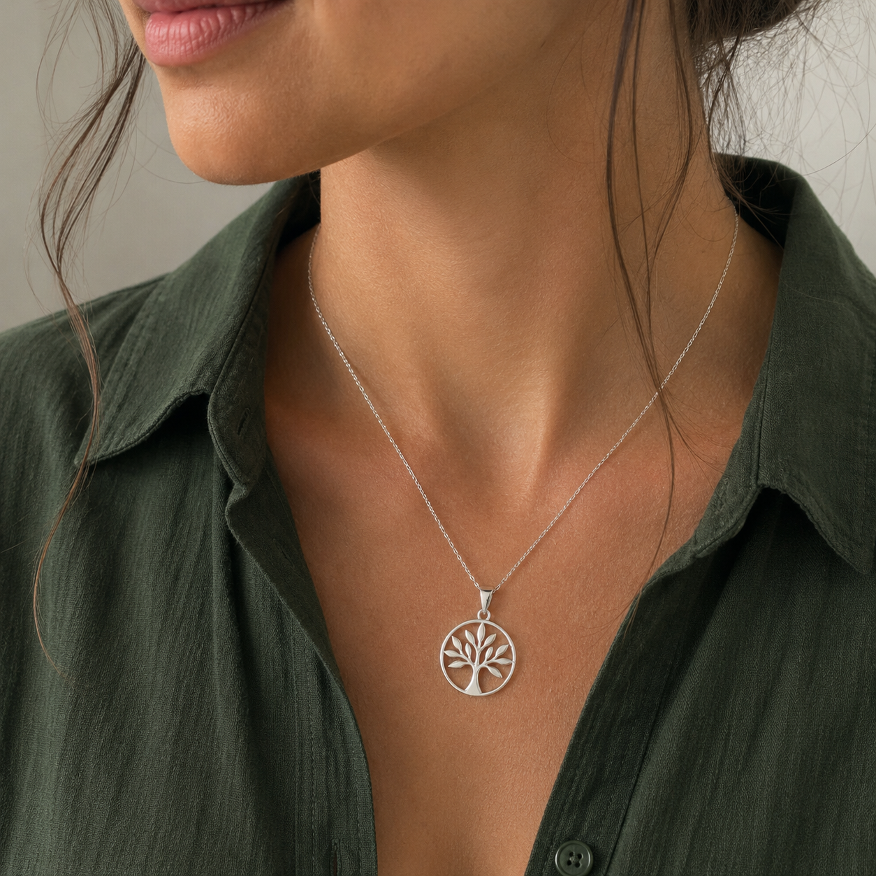 A close-up of a woman wearing a delicate silver tree of life pendant necklace, styled with an olive green collared shirt.
