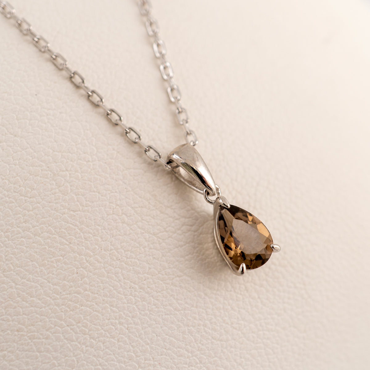 Angled view of a smoky quartz teardrop necklace draped over a white cushion, showing the pendant facing right and its translucent brown gemstone shades.