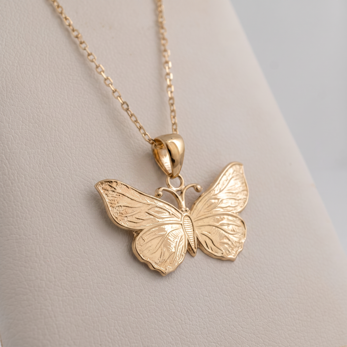 A gold butterfly pendant necklace displayed on a light beige jewelry stand against a soft gray background.