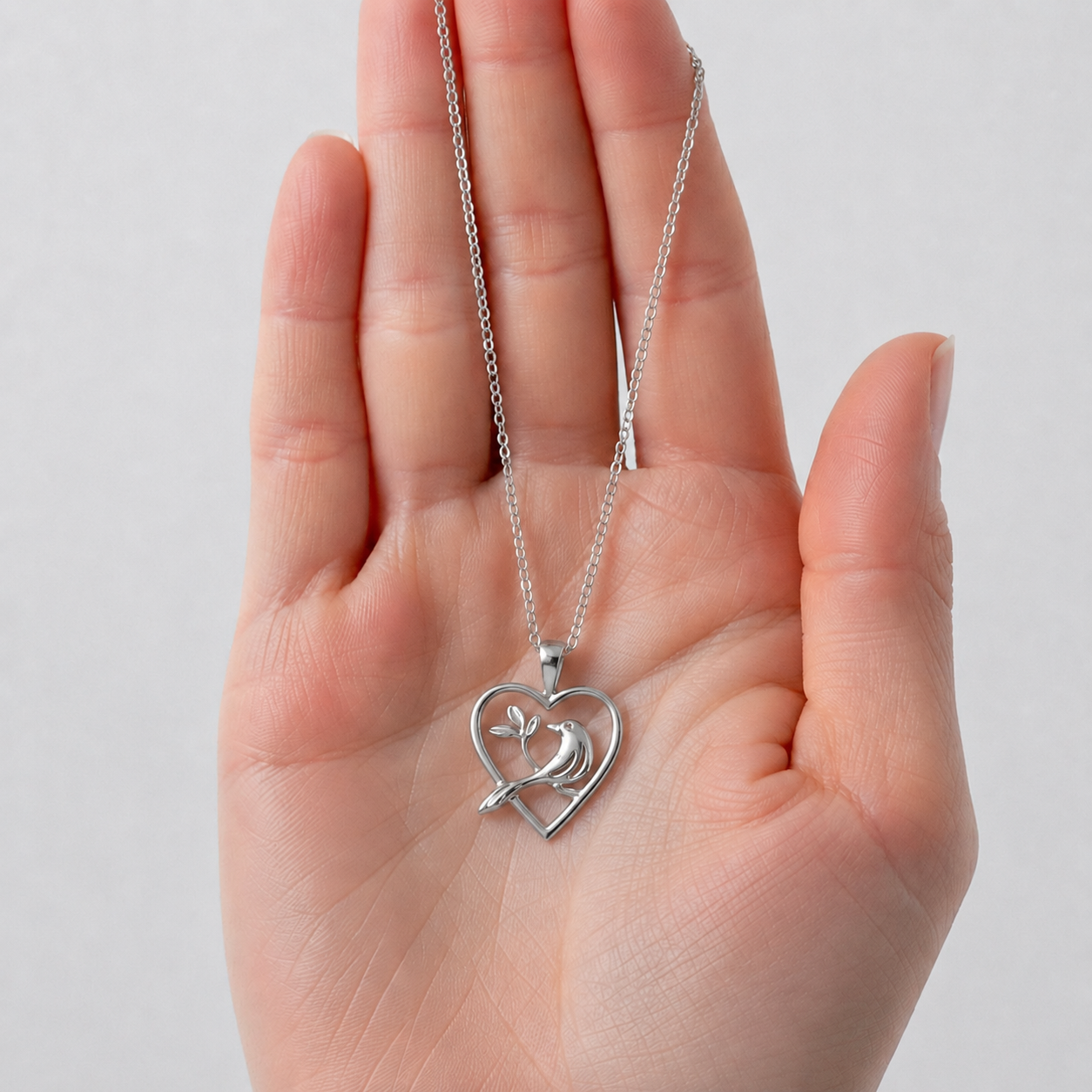 A silver heart-shaped pendant necklace with a dove in the center, held in a person's open hand against a light gray background.