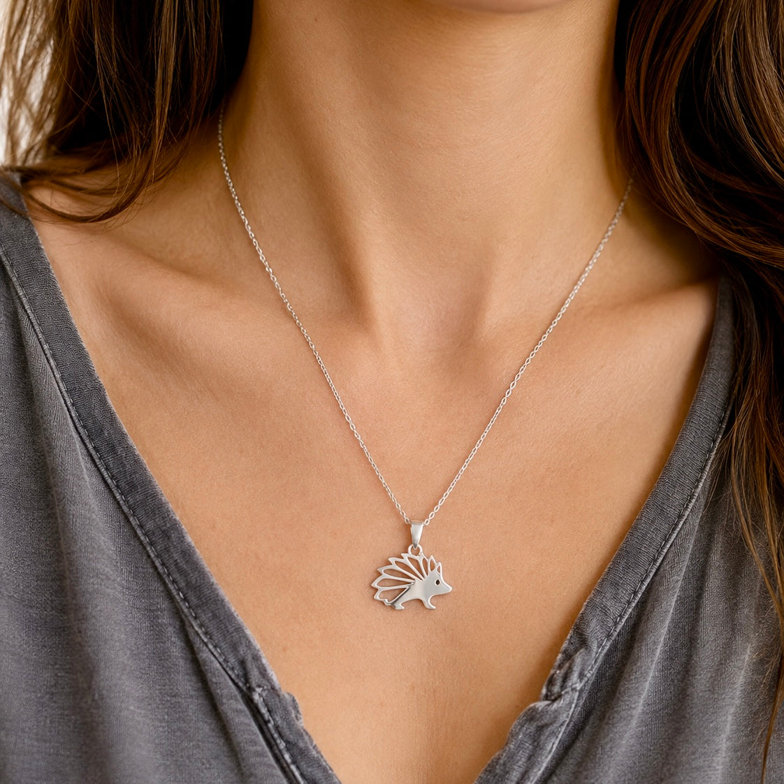 A close-up of a person wearing a delicate silver chain necklace with a small hedgehog pendant, styled with a gray V-neck top.