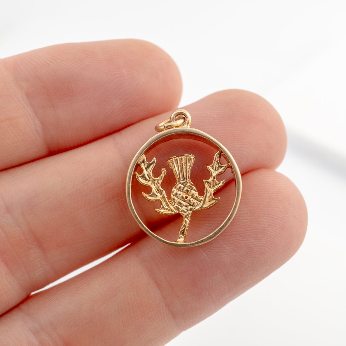 A gold-plated Scottish thistle pendant in a circular openwork design, held on a person's fingers to show its small scale.