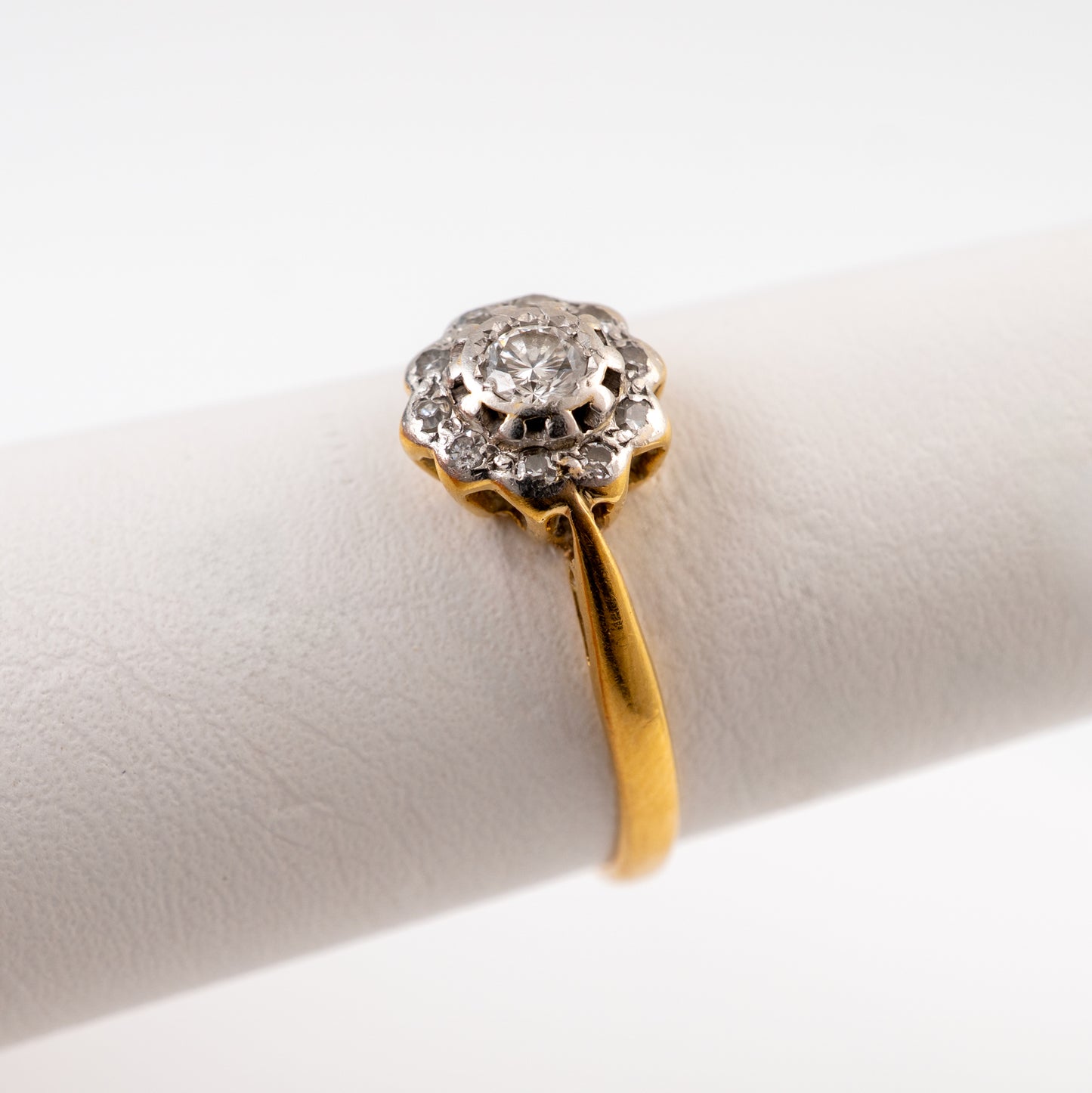 A vintage diamond ring with a yellow gold band and a silver-toned floral halo holding a round clear gemstone, displayed on a white ring stand.