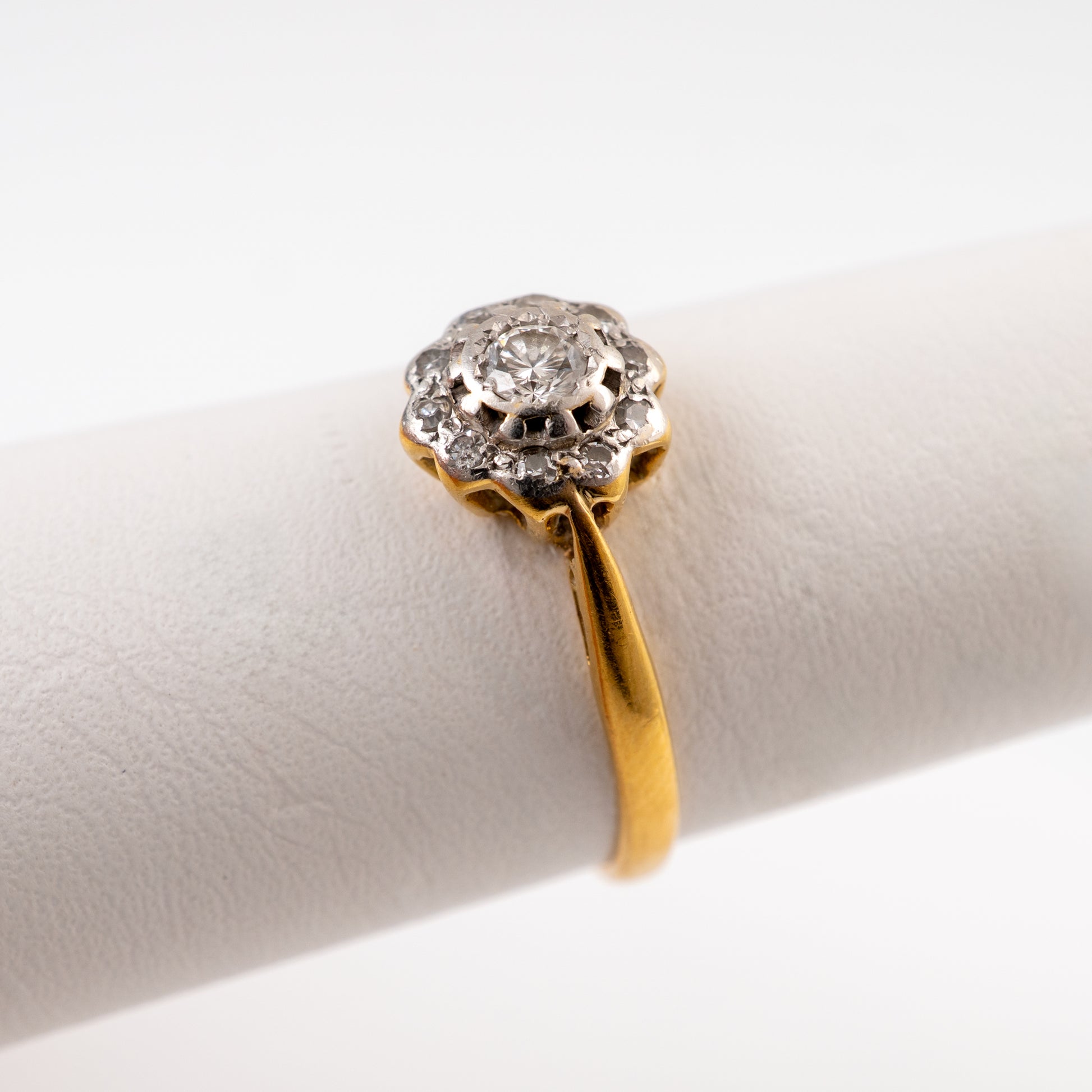 A vintage diamond ring with a yellow gold band and a silver-toned floral halo holding a round clear gemstone, displayed on a white ring stand.