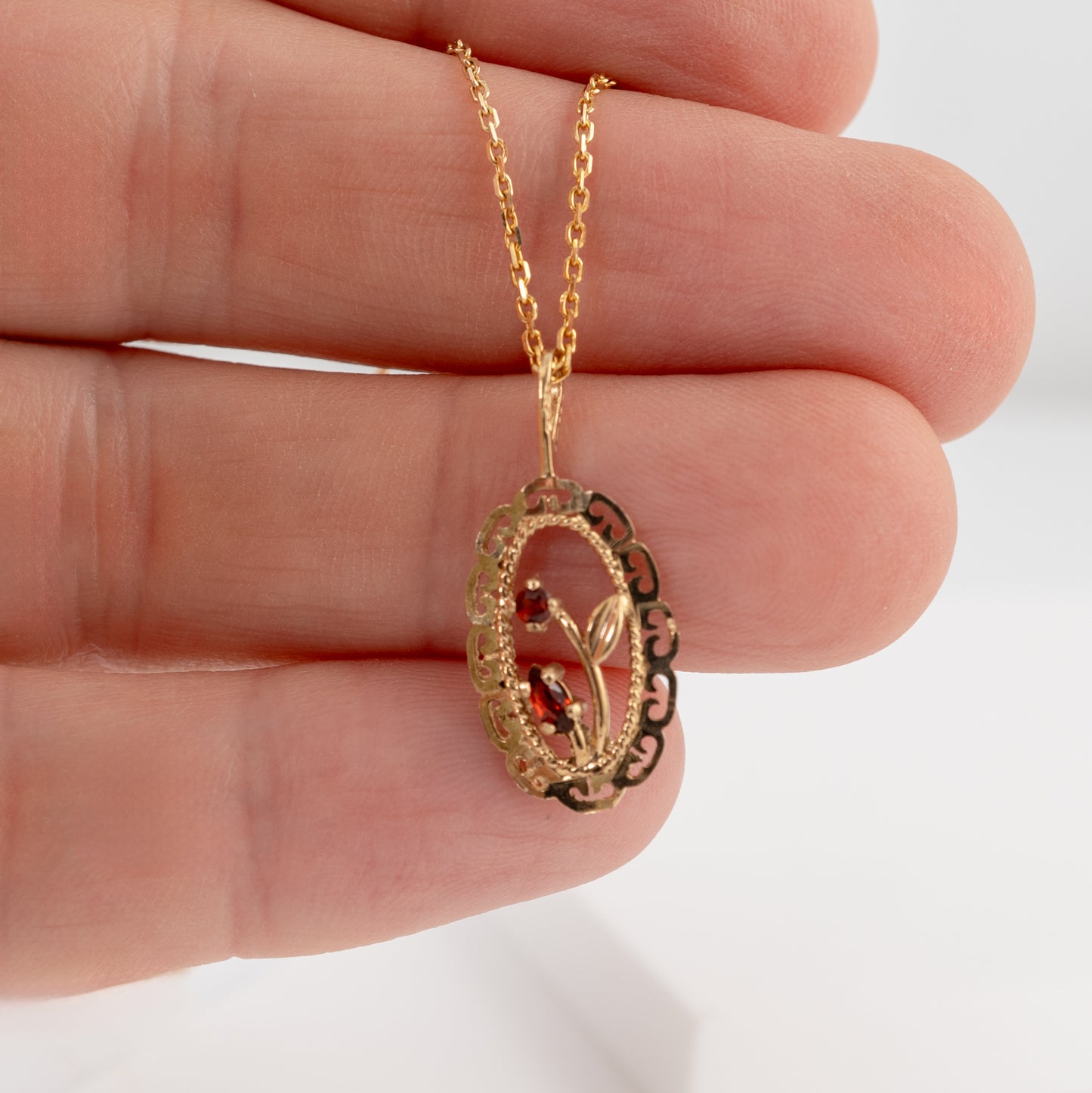 A gold pendant necklace with a delicate floral design and small amber-red stones, held in a person's hand against a white background.