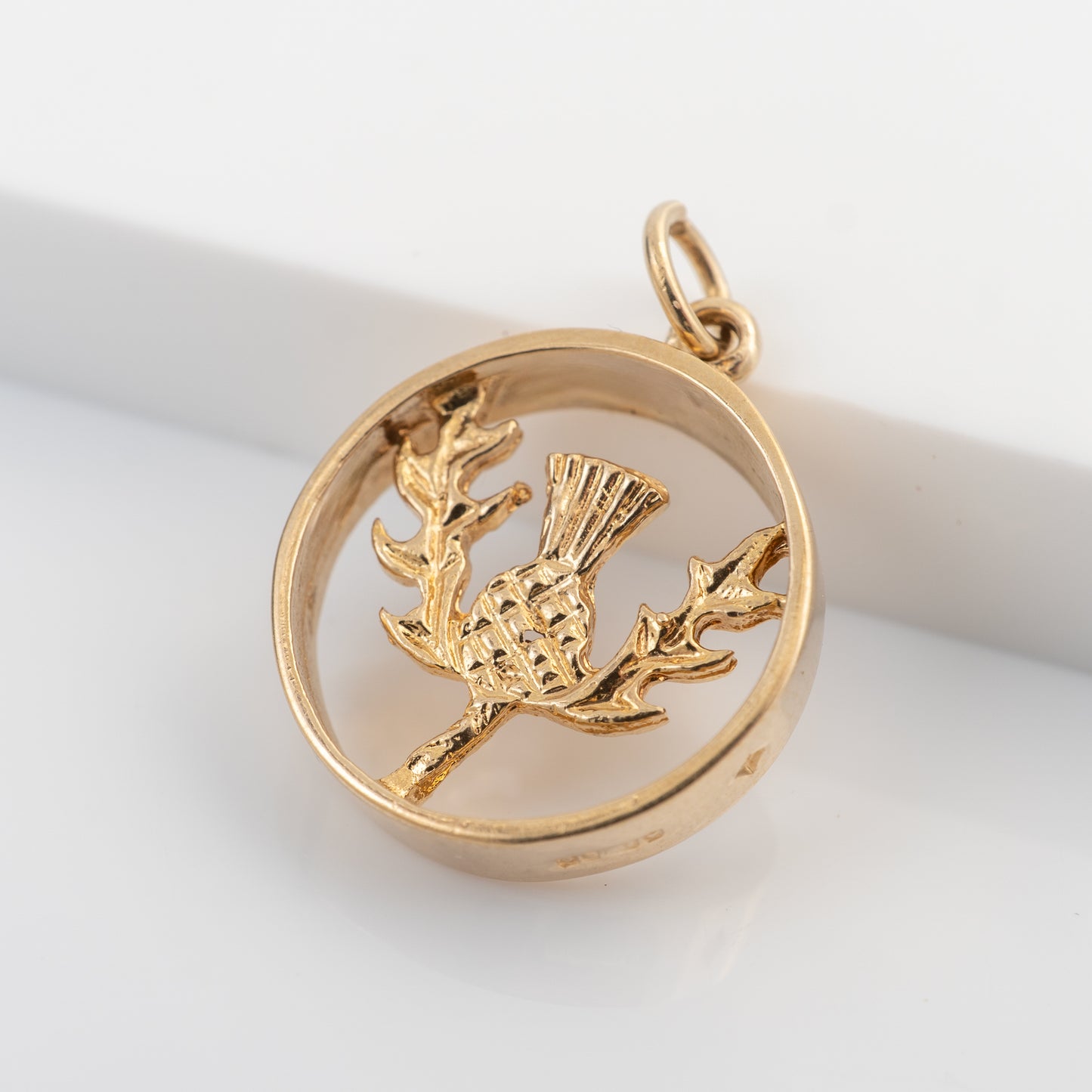 A gold-toned circular pendant charm featuring a raised bird perched on a branch, photographed on a clean white background.