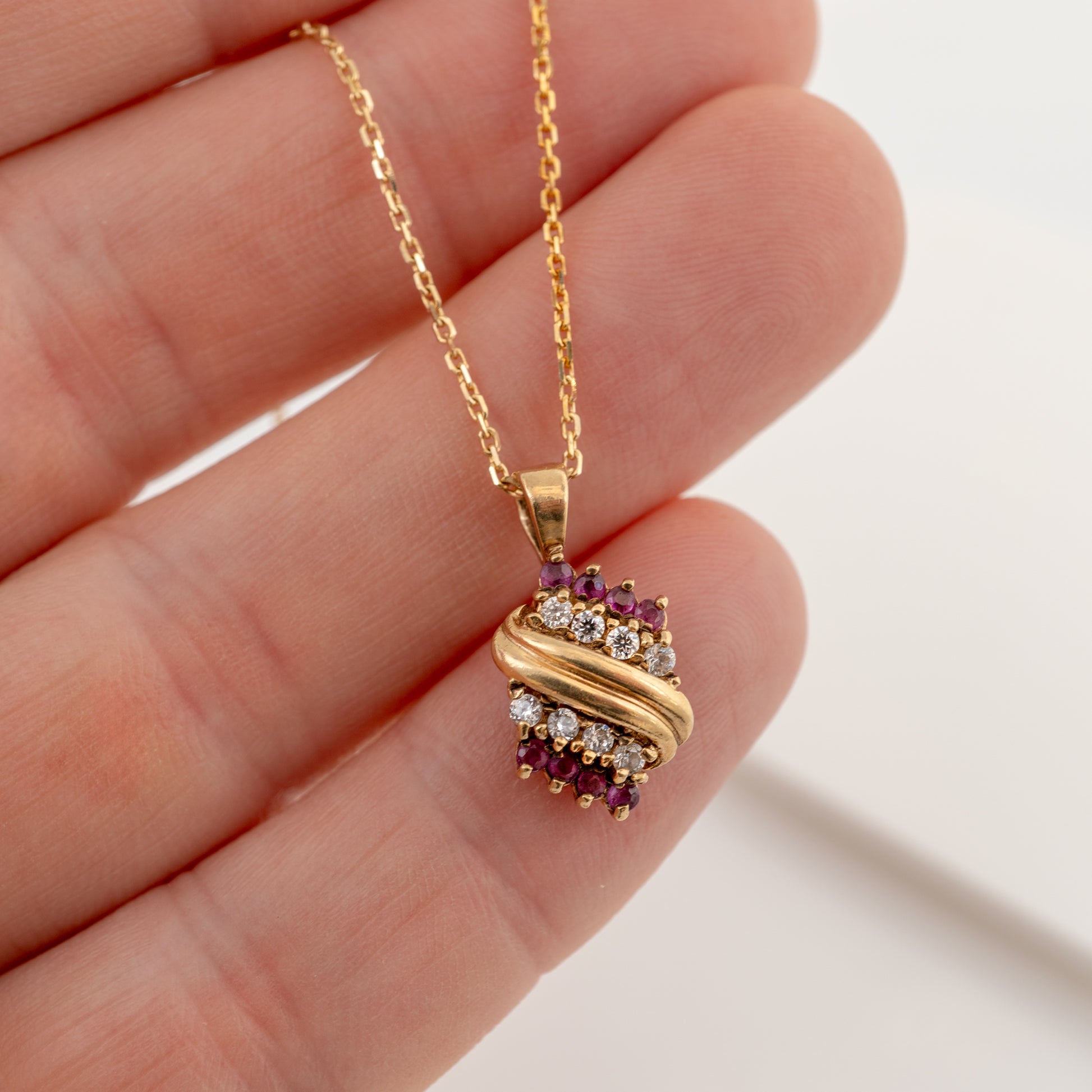 A gold pendant necklace with a twisted wrap design, accented by small purple gemstones and clear stones, held in a person's hand against a light background.