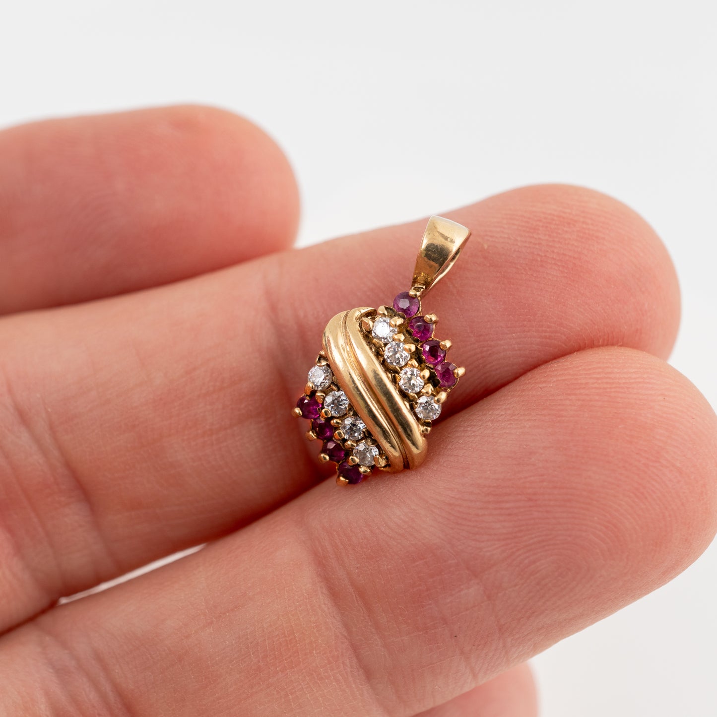 A gold pendant with twisted bands and rows of clear and dark red gemstones, shown resting on a person's fingers against a white background.