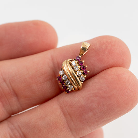 A gold pendant with twisted bands and rows of clear and dark red gemstones, shown resting on a person's fingers against a white background.