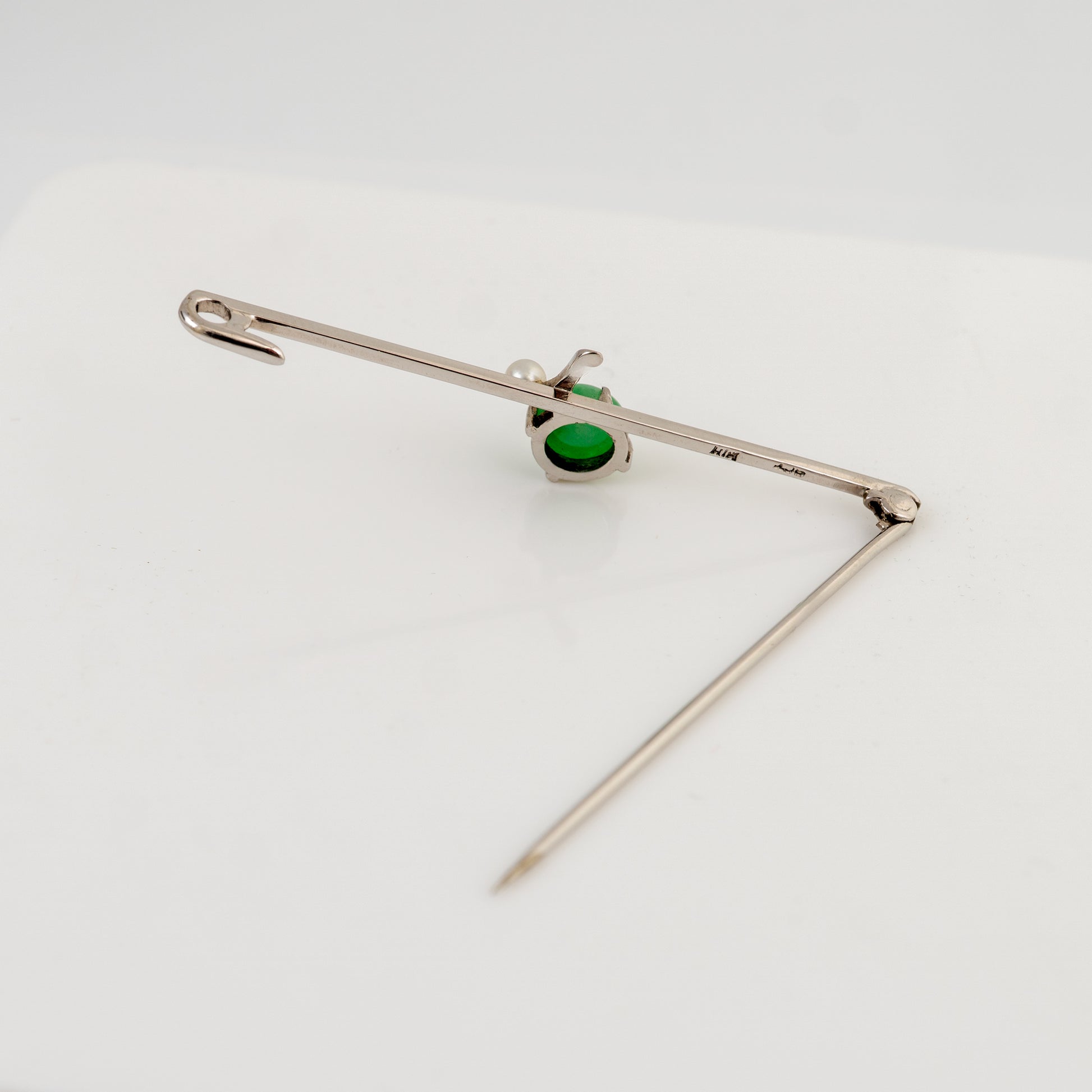 A silver-toned safety pin brooch with a small green oval stone and a tiny white pearl accent, displayed on a clean white surface.