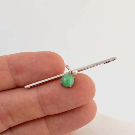 A white gold bar pin with a round green jadeite gemstone and a small pearl accent, held between fingers to show scale.