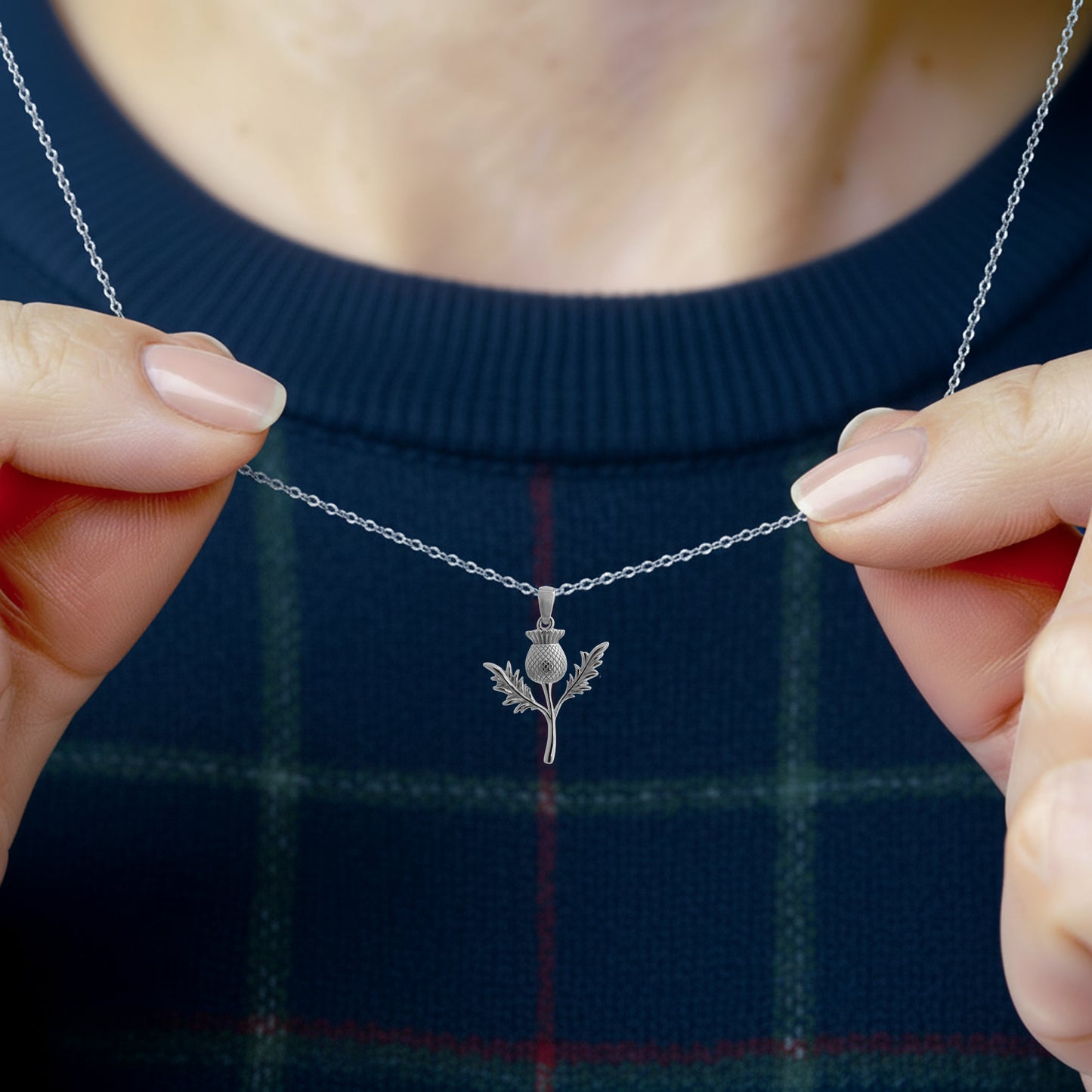 Silver thistle necklace held by hands against a dark blue plaid background