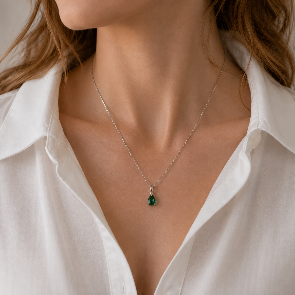 A close-up of a woman wearing a delicate silver chain necklace with a small teardrop-shaped emerald gemstone pendant, styled with a white collared blouse.