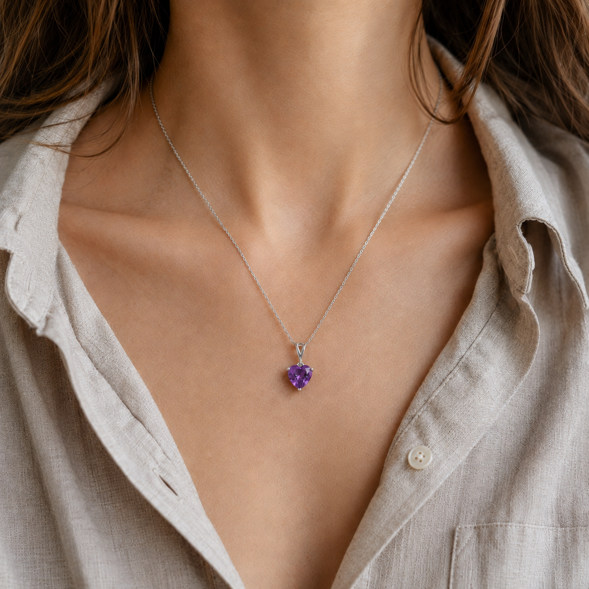 A close-up of a person wearing a delicate silver chain necklace with a small heart-shaped purple gemstone pendant, styled with a light beige linen shirt.