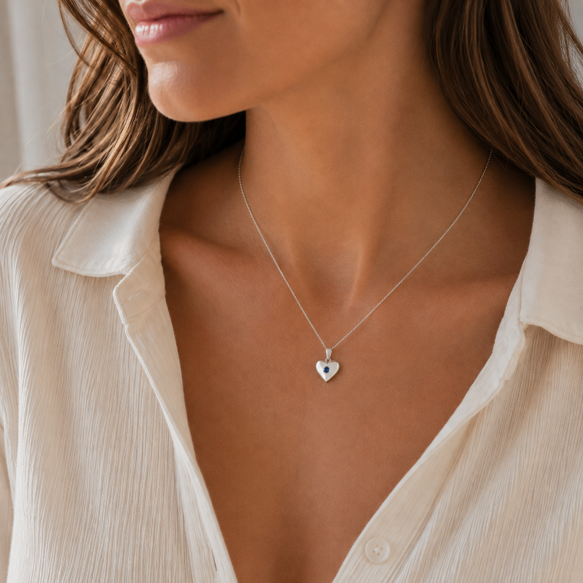 A close-up of a woman wearing a delicate silver heart pendant necklace with a small dark blue stone in the center, styled with a white collared blouse.